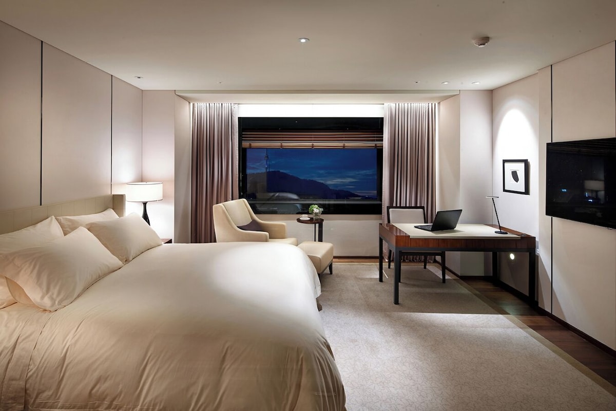 A hotel room with a large bed, a desk with a laptop, a chair, a TV, and a window with curtains.