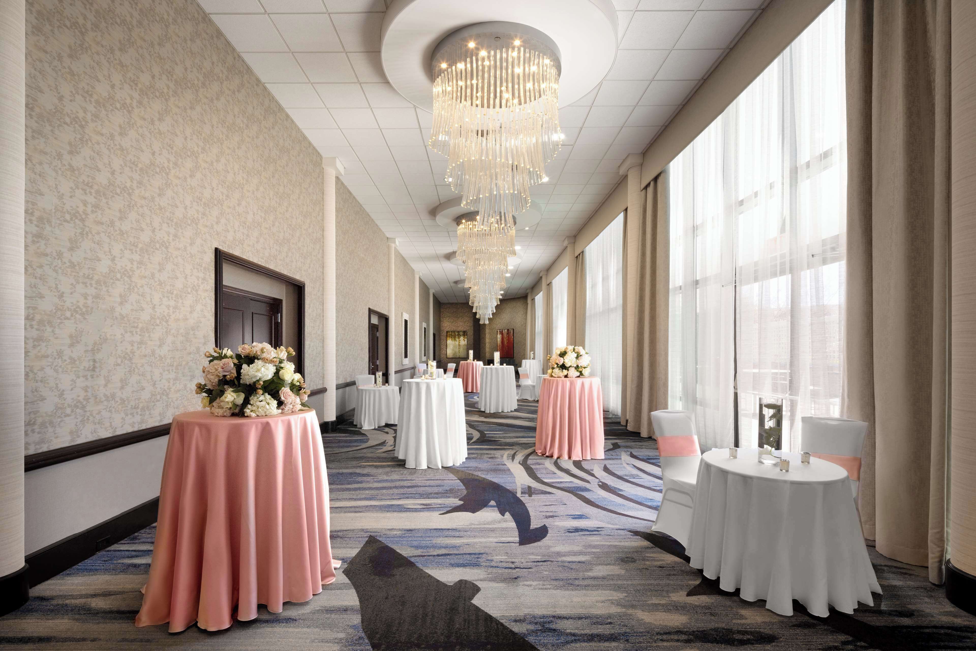 A hotel corridor with round tables, floral centerpieces, and a large chandelier.