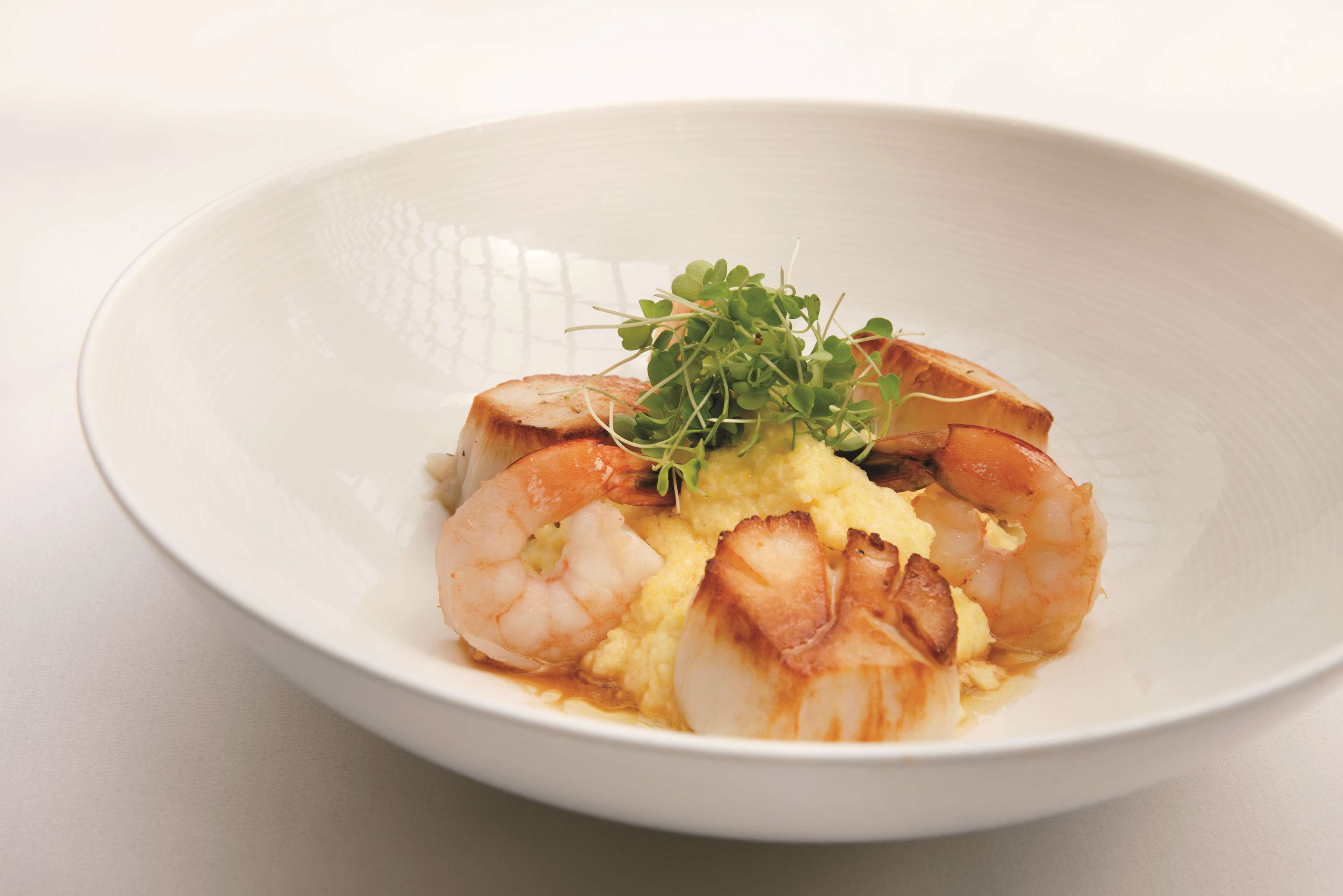 A white bowl containing a seafood dish with shrimp and scallops, garnished with microgreens.