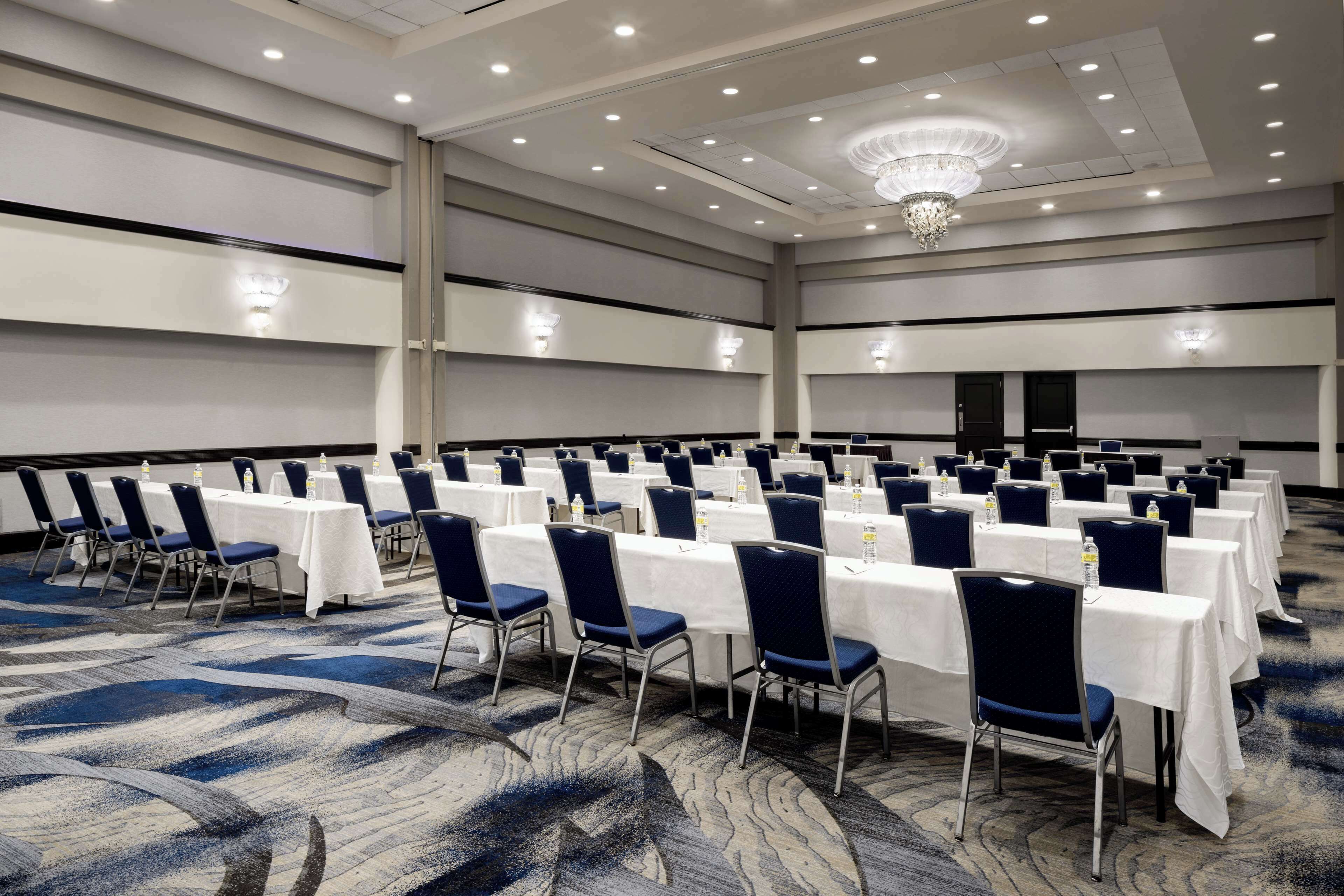 Een ruime congreszaal met rijen tafels en stoelen, voorzien van waterflesjes en een kroonluchter.