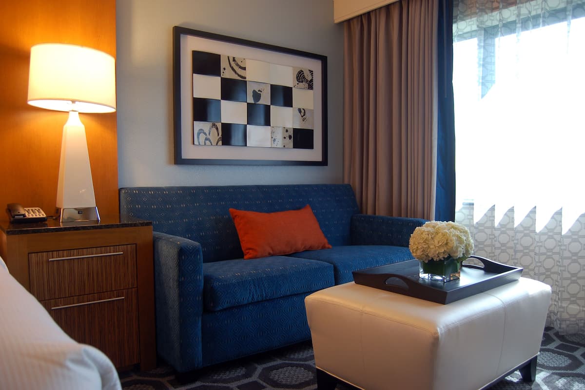 Habitación de hotel con sofá azul, reposapiés blanco, escritorio con lámpara y una obra de arte abstracta enmarcada en la pared.