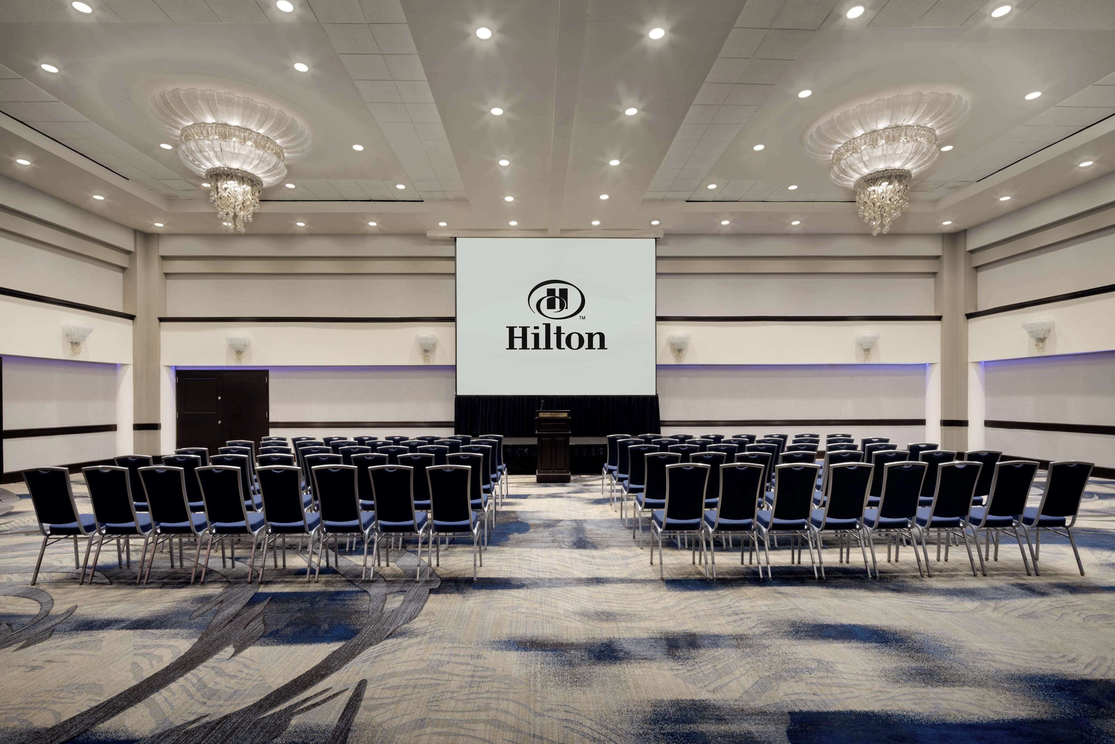 Un salón de conferencias con filas de sillas, un atril y una pantalla grande mostrando el logotipo de Hilton.