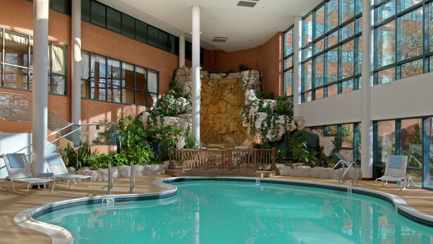 An indoor swimming pool with a stone wall and plants, surrounded by windows and lounge chairs. - Hilton Long Island/Huntington (Melville)