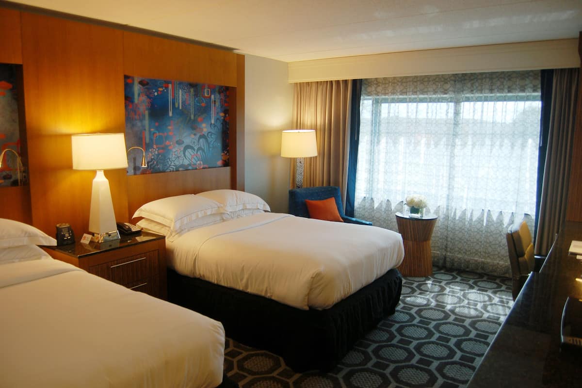 Habitación de hotel con dos camas, un cuadro grande, un escritorio con lámpara y vistas al exterior a través de una ventana con cortinas.