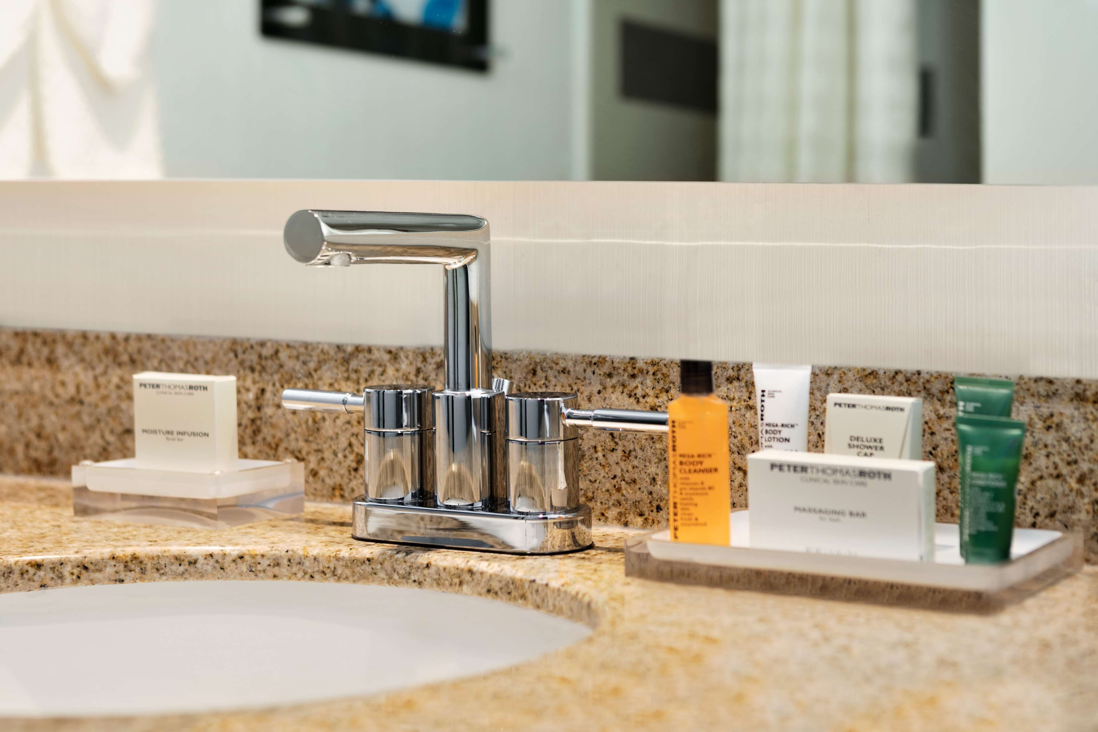 A bathroom vanity with a chrome faucet, a sink, and a mirror. There are toiletries on the counter, including a bottle of lotion and a box labeled "Peter Thomas Roth".