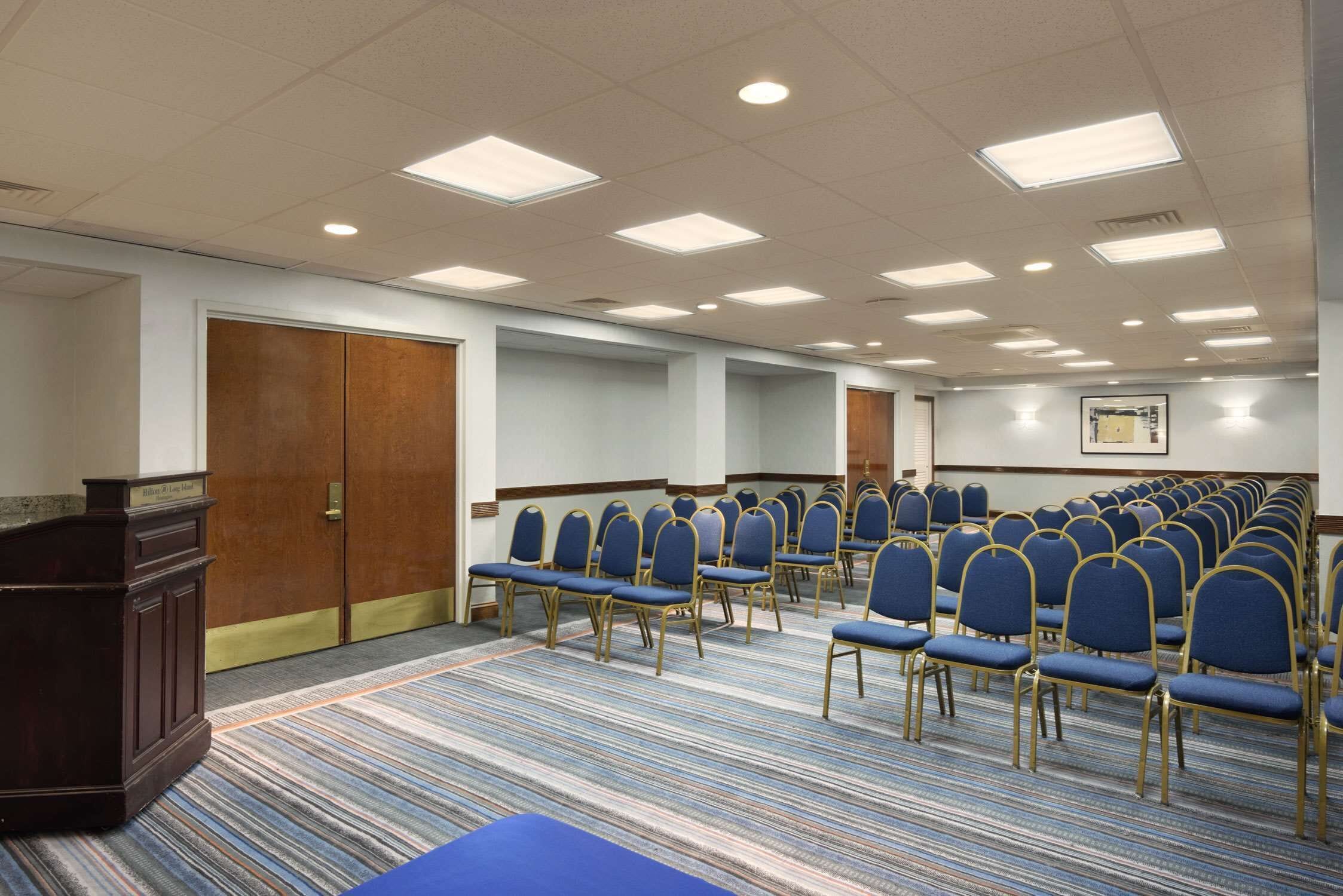 A conference room with rows of blue chairs, a podium, and a carpeted floor.