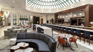 A modern hotel lobby with a curved seating area, a bar with a long counter, and a large glass ceiling. - Hilton Long Island/Huntington (Melville)