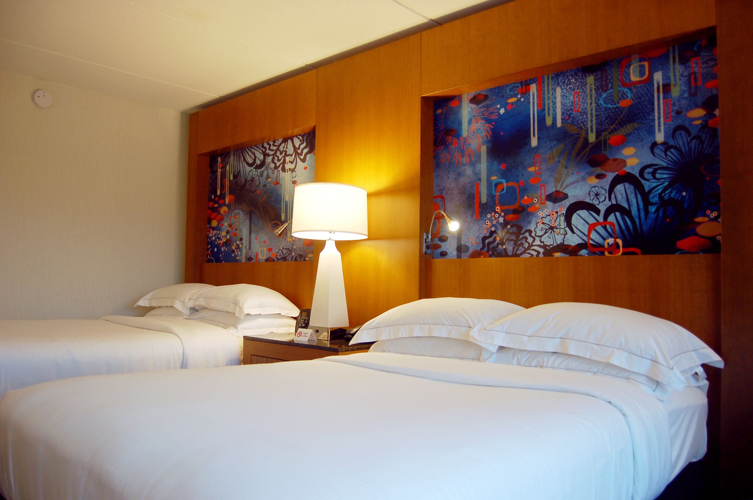 A hotel room with two beds, a nightstand with a lamp, and two wall paintings.