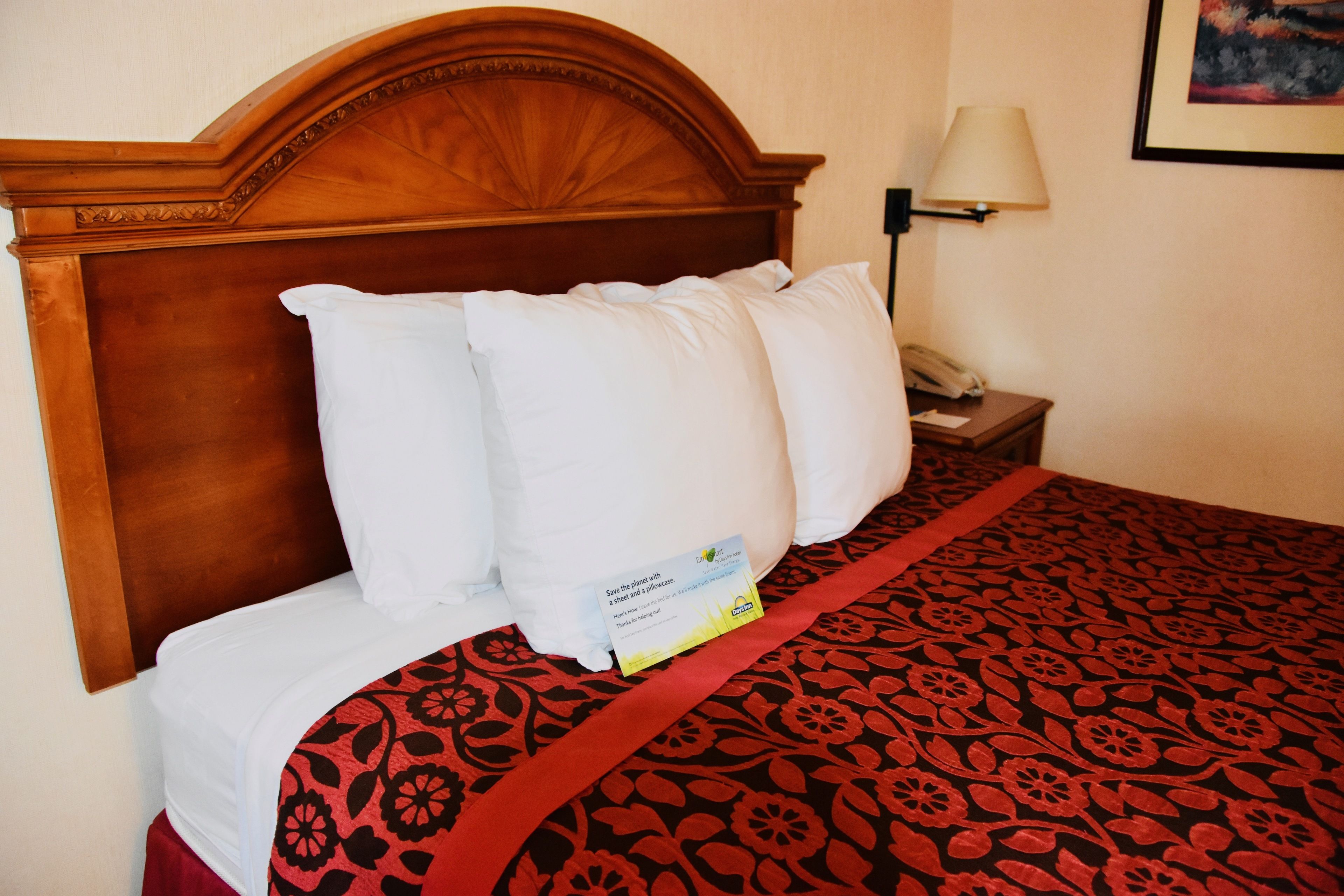 A neatly made bed with a red and black patterned bedspread, a wooden headboard, and a bedside table with a lamp and a phone.