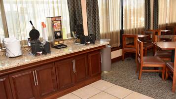 A kitchen area with a coffee machine, a waffle maker, and a refrigerator. There are wooden chairs and a table in the dining area.