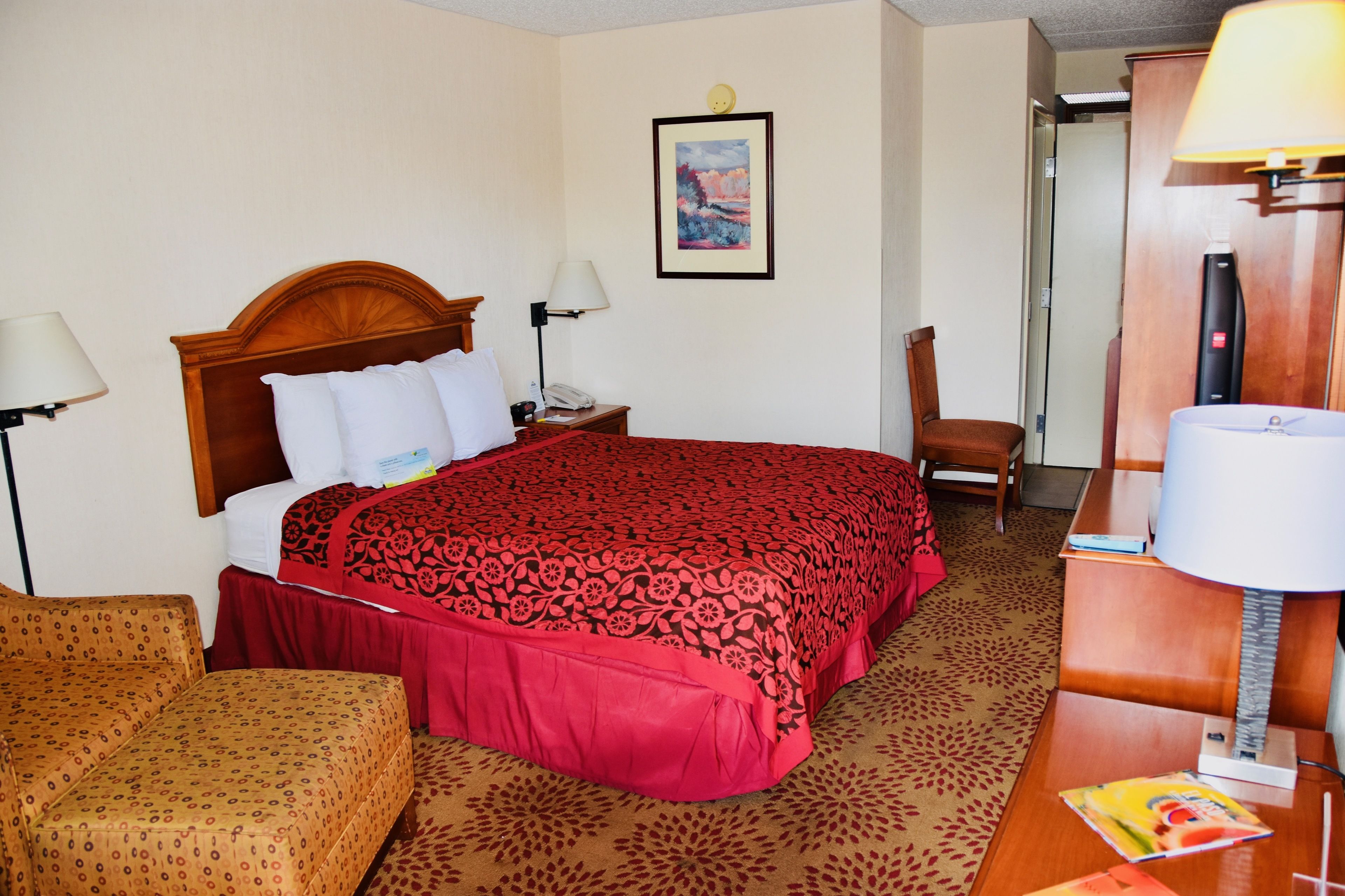 A hotel room with a large bed, a wooden headboard, two bedside lamps, a chair, a desk with a lamp, and a framed picture on the wall.
