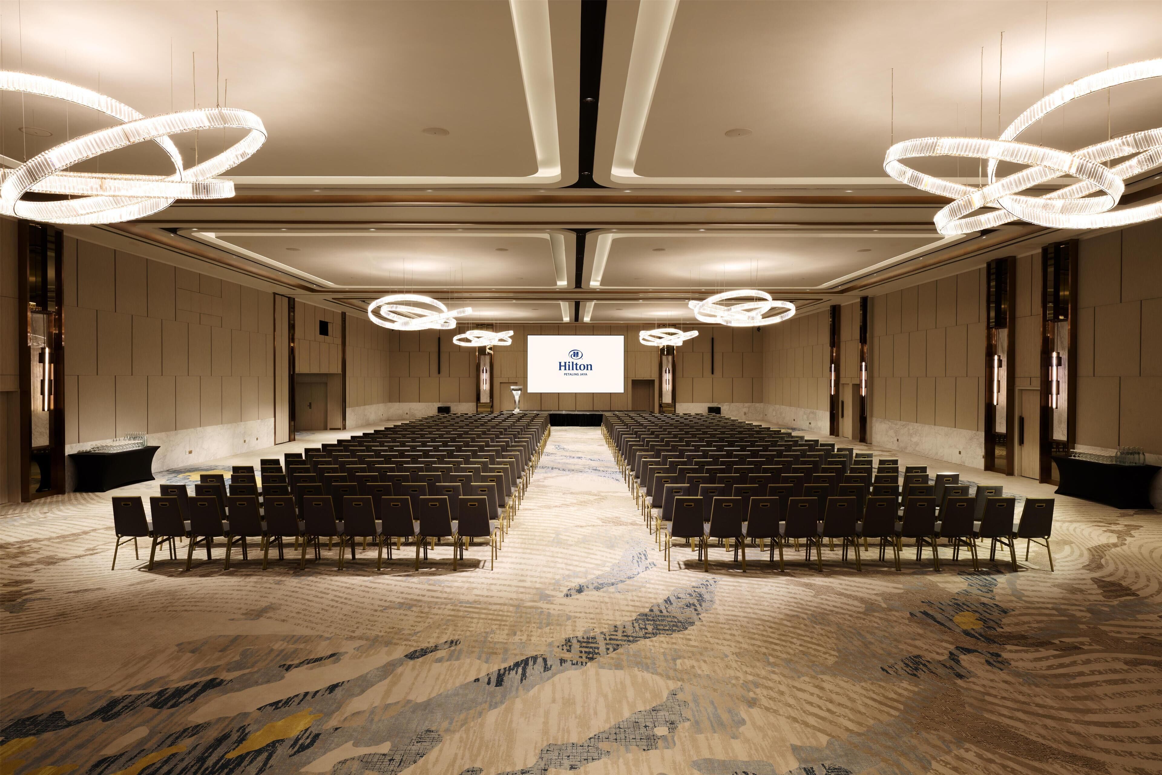 A large conference room with rows of chairs, a central screen, and modern lighting fixtures.