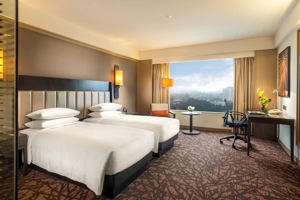 A hotel room with two beds, a desk, a chair, a small table, and a view of the city.