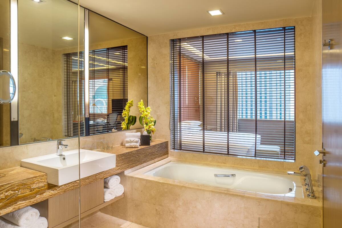 A modern bathroom with a large mirror, two sinks, a bathtub, and a window with blinds.