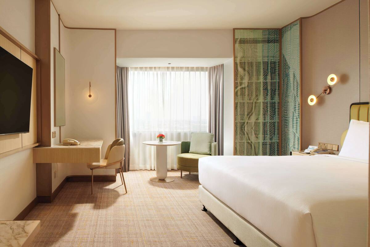 A hotel room with a large bed, a desk with a telephone, a chair, a small round table with a flower, and a TV mounted on the wall.