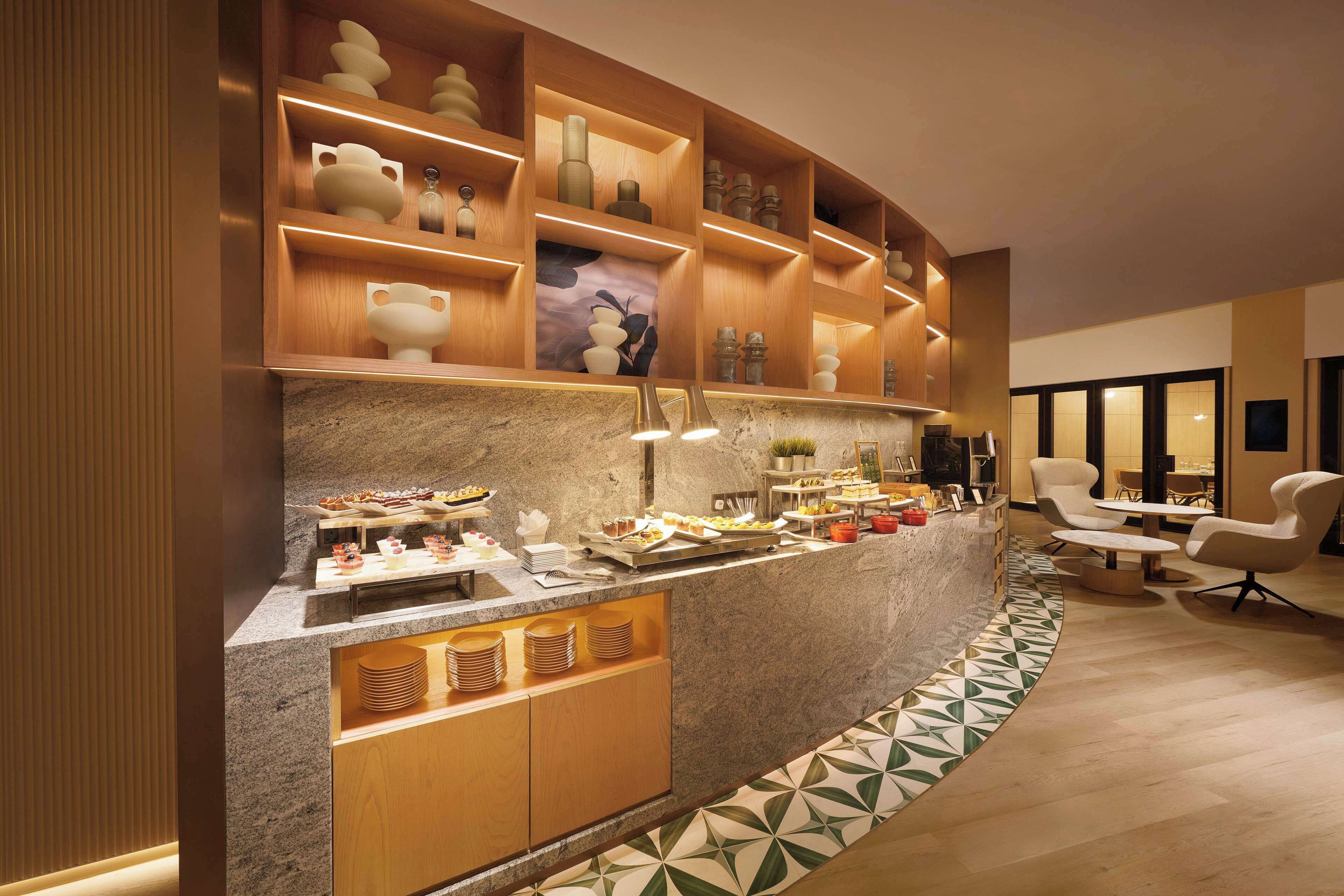 A modern hotel lobby with a curved reception desk, wooden shelving with decorative items, and a seating area with white chairs.