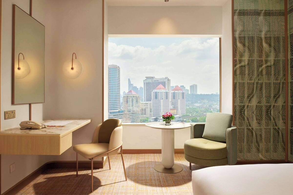 A hotel room with a desk, two chairs, a bed, a round table with a vase, and a large window with a city view.