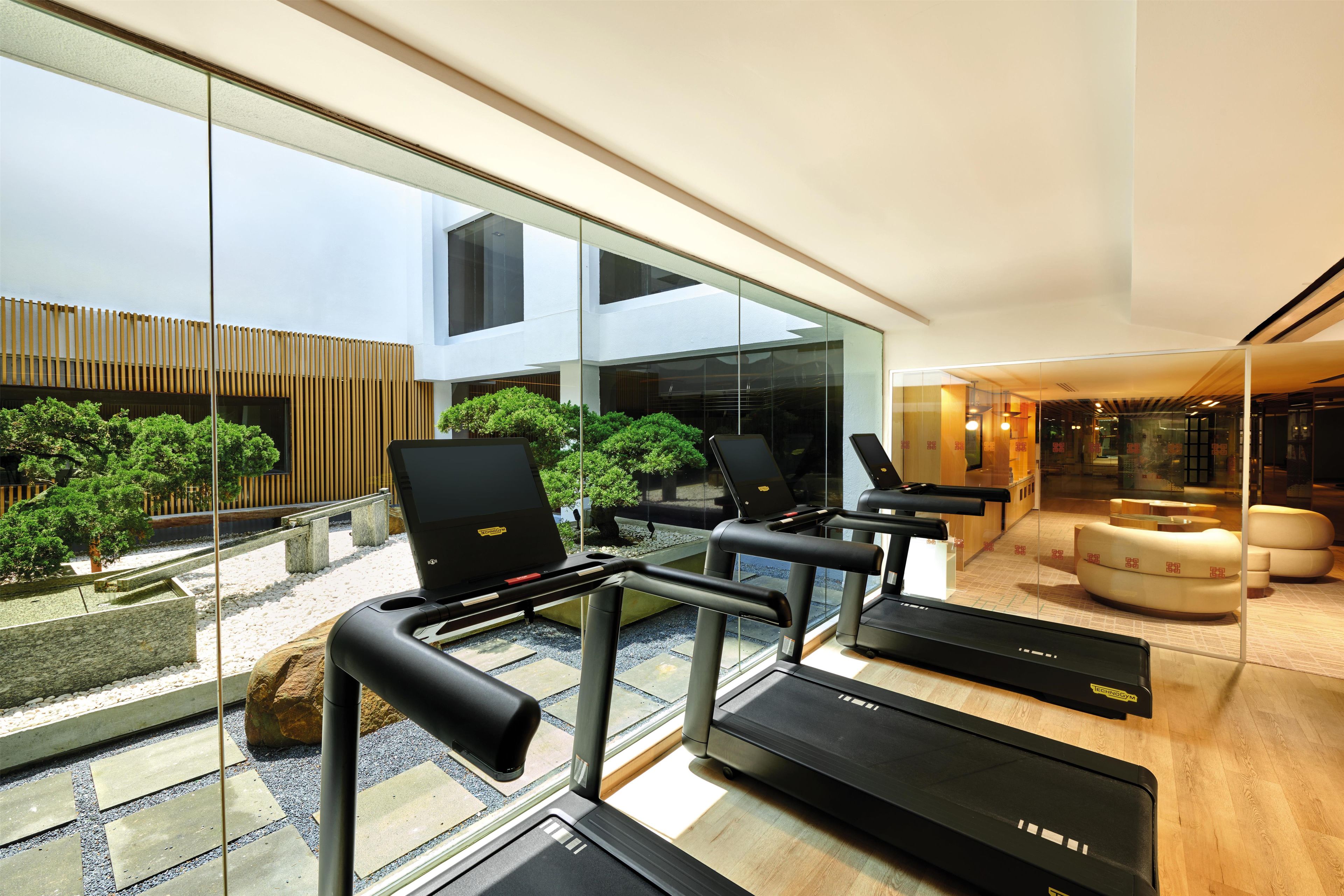 A modern gym with exercise equipment, a wooden floor, and a glass wall overlooking a courtyard with trees.