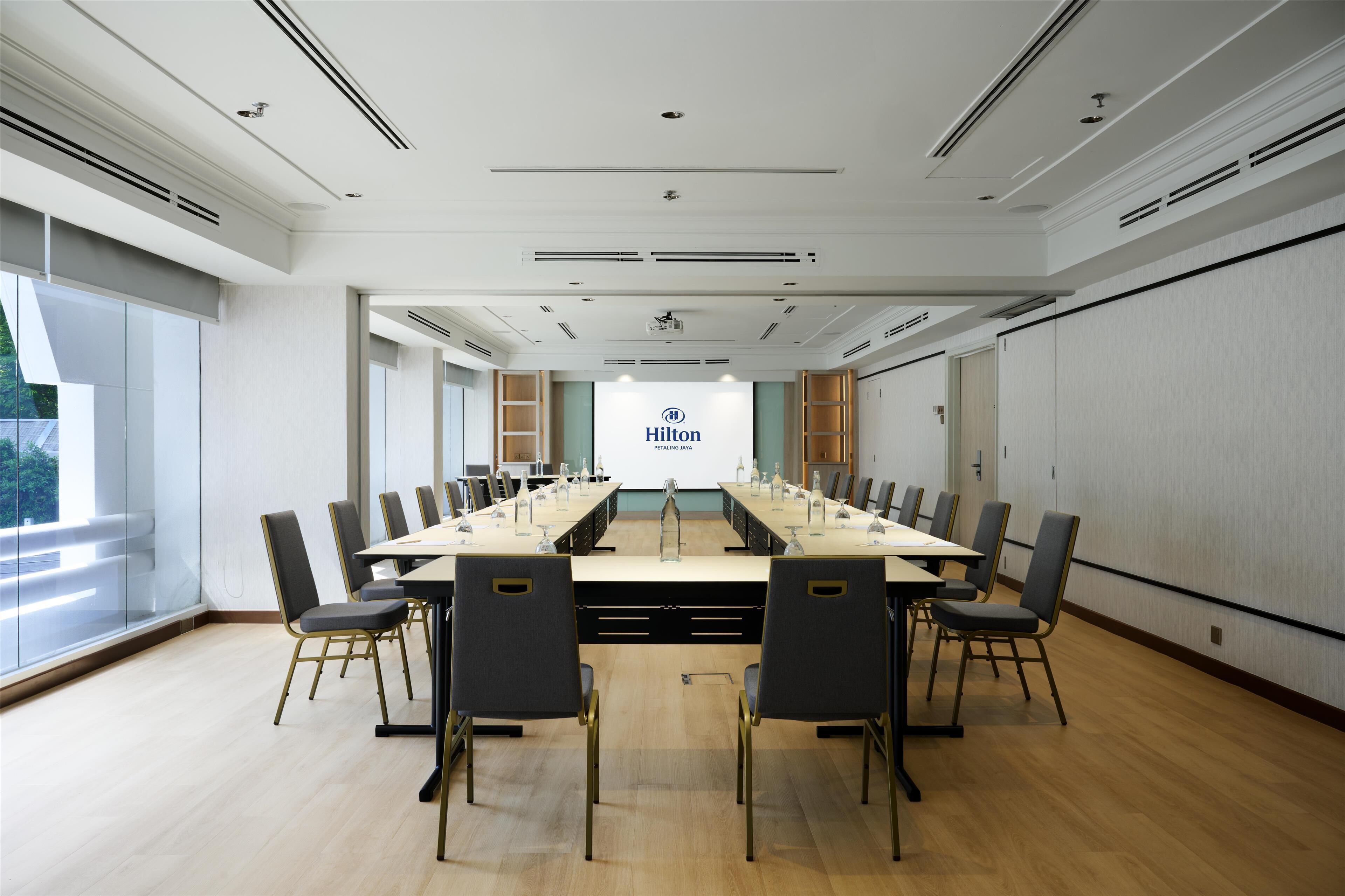A conference room with a long table, chairs, a projector screen, and a view of the outdoors.