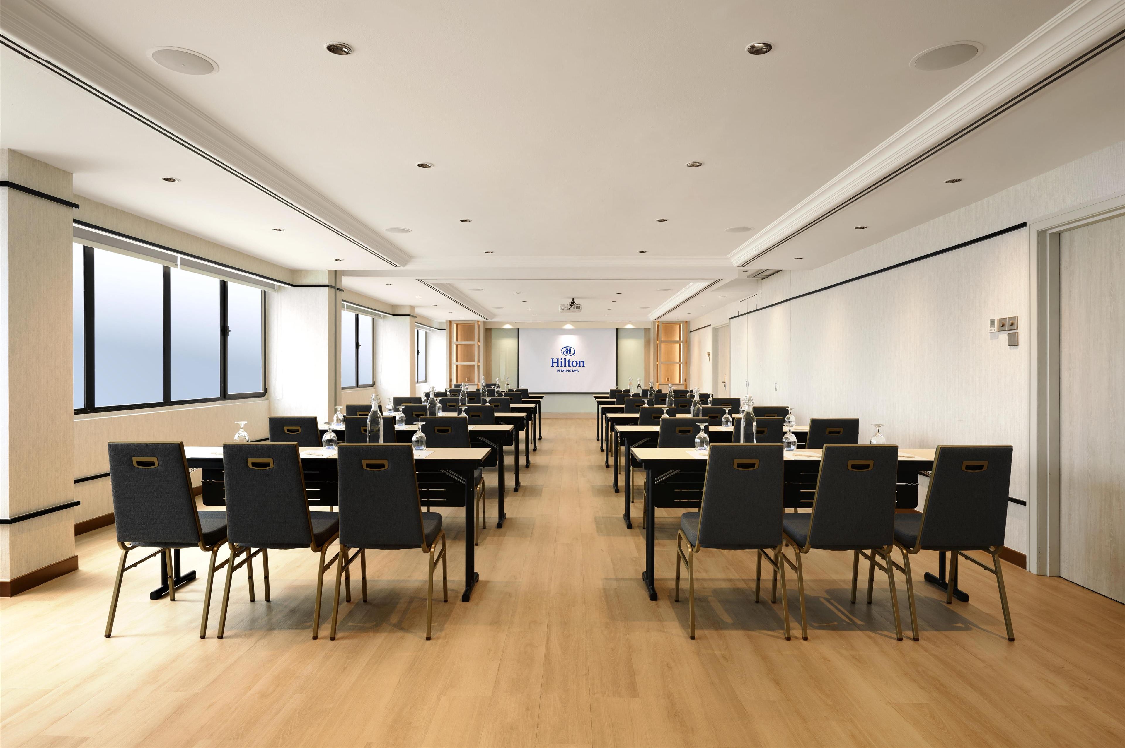 A conference room with multiple tables and chairs, a projector screen, and a large window on one side.
