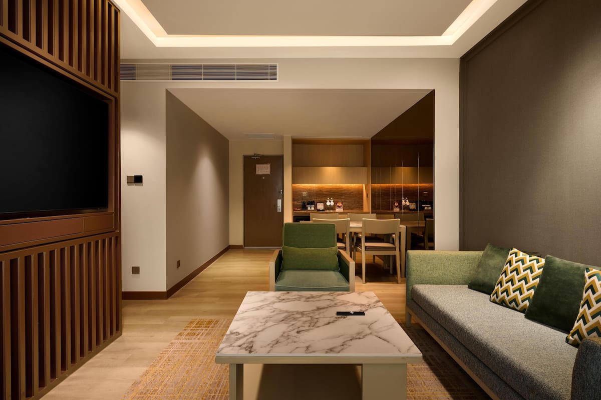 A modern living room with a sofa, a coffee table, and a TV. There is a dining area with chairs and a table in the background.