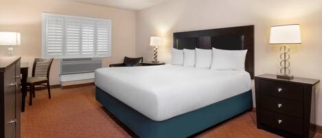A hotel room with a large bed, a desk, a chair, a nightstand, and a lamp.