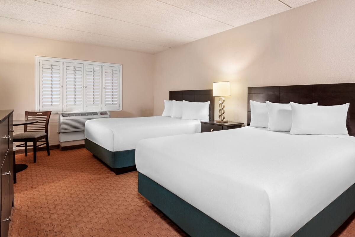 Une chambre d’hôtel avec deux lits, un bureau, une chaise et une fenêtre avec des stores.
