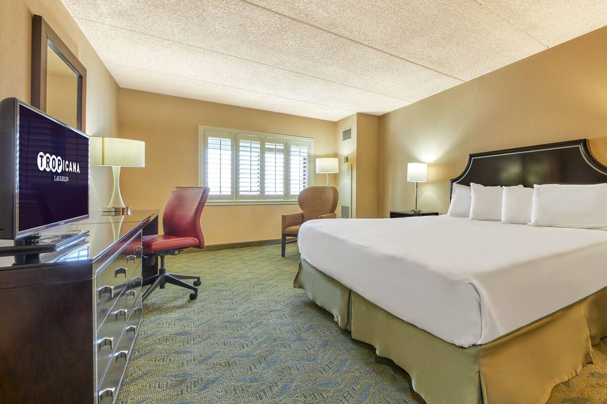 Une chambre d’hôtel avec un grand lit, un bureau avec une télévision, deux chaises, une fenêtre avec des stores et un sol recouvert de moquette.