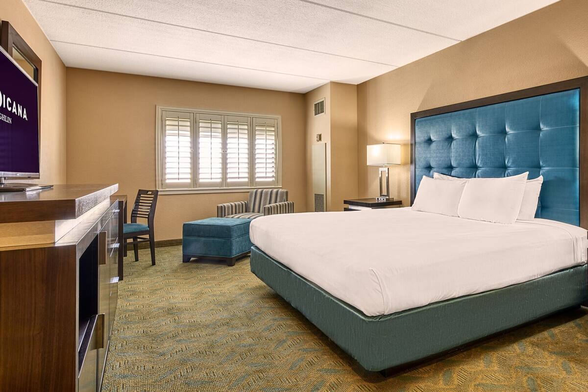 Une chambre d’hôtel avec un grand lit, un bureau, une chaise, une lampe et une fenêtre avec des stores.
