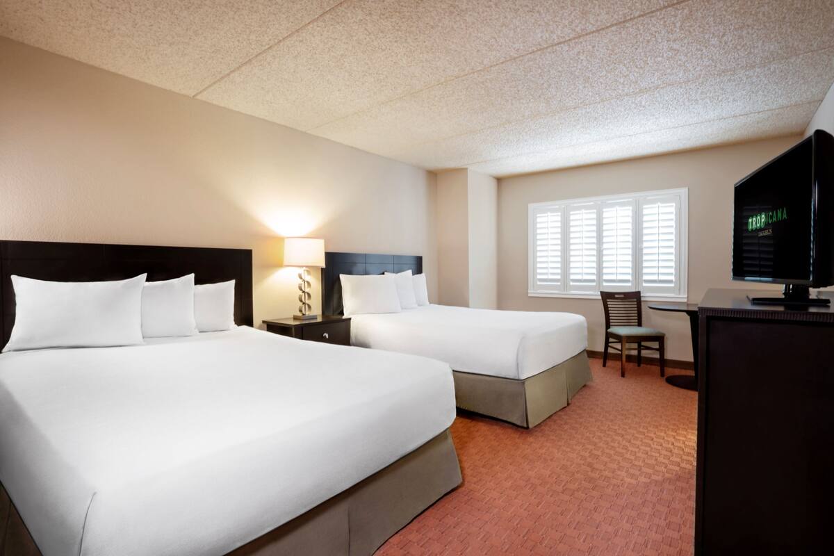 Une chambre d’hôtel avec deux lits, un bureau, une chaise, une télévision et une fenêtre avec des stores.