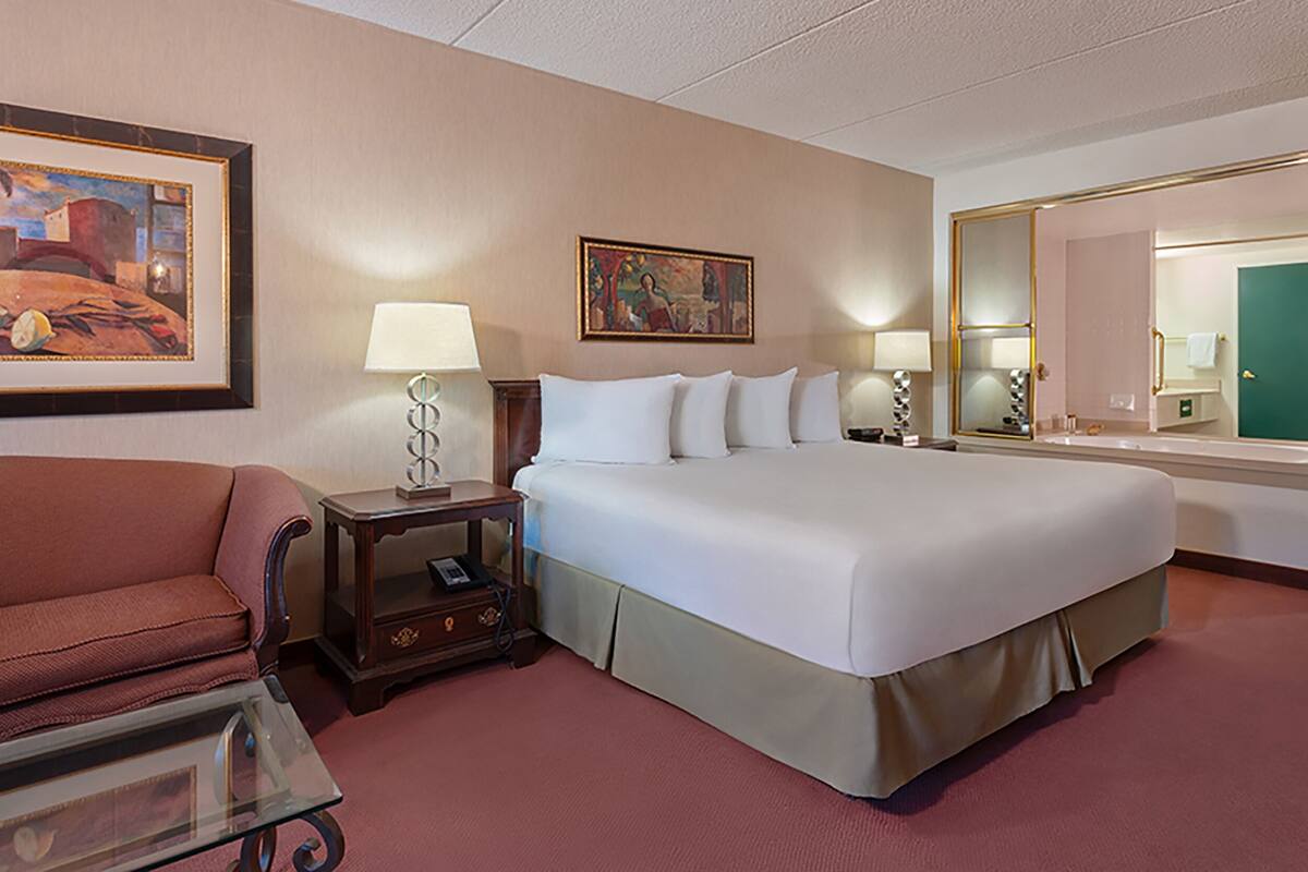 Une chambre d’hôtel avec un grand lit, un bureau avec une lampe, une chaise, une petite table, un tableau et un miroir.