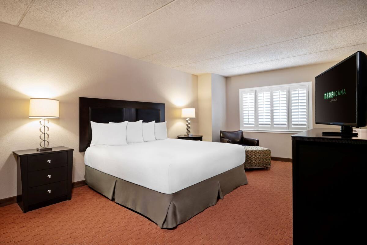 Une chambre d’hôtel avec un grand lit, deux tables de chevet avec des lampes, une télévision et une fenêtre avec des stores.