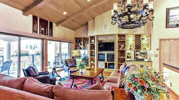 A spacious living area with a wooden ceiling, a chandelier, a fireplace, and a large window overlooking a balcony.