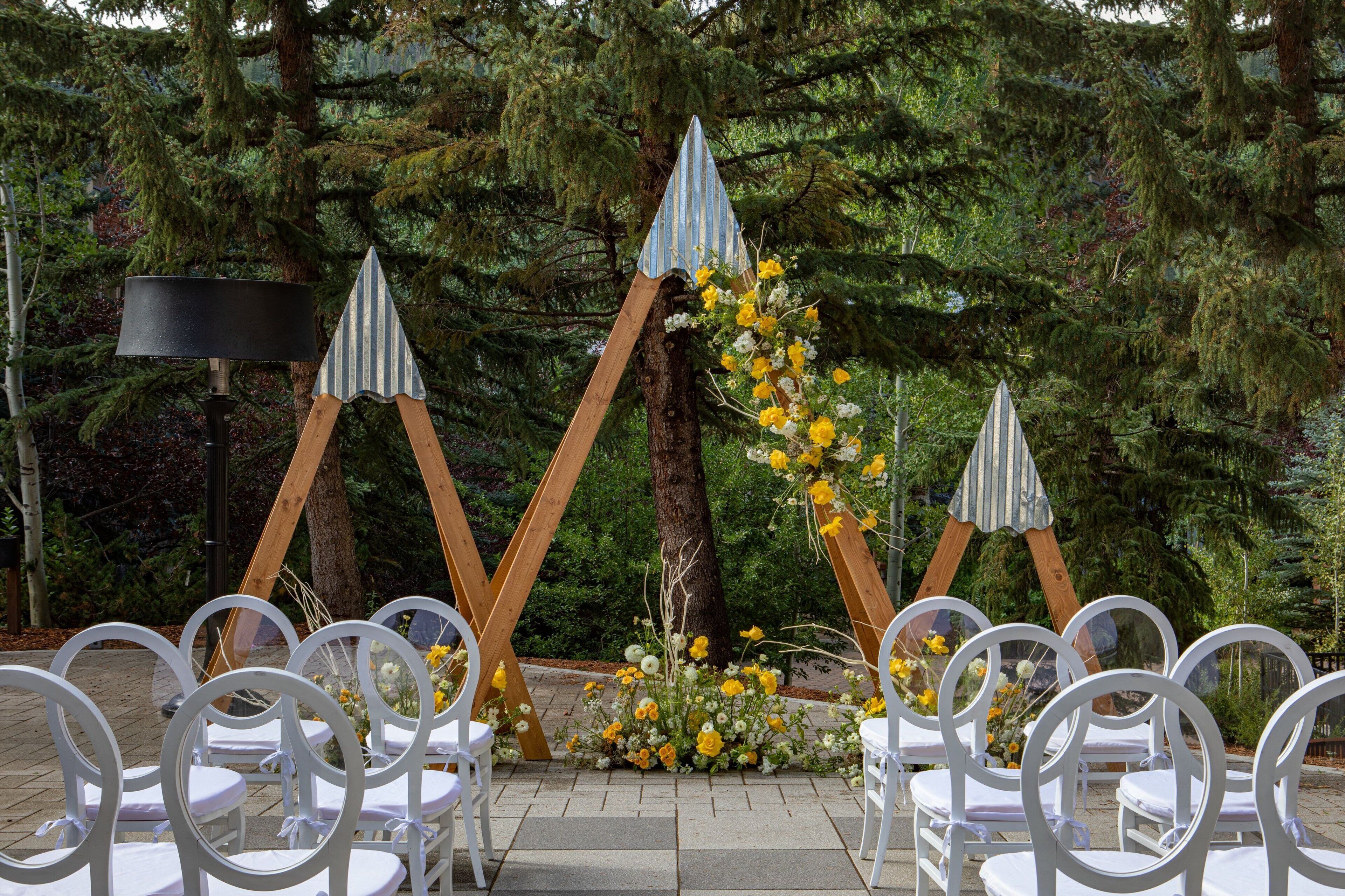 Montaje para una boda al aire libre con sillas blancas, un arco de madera y arreglos florales.