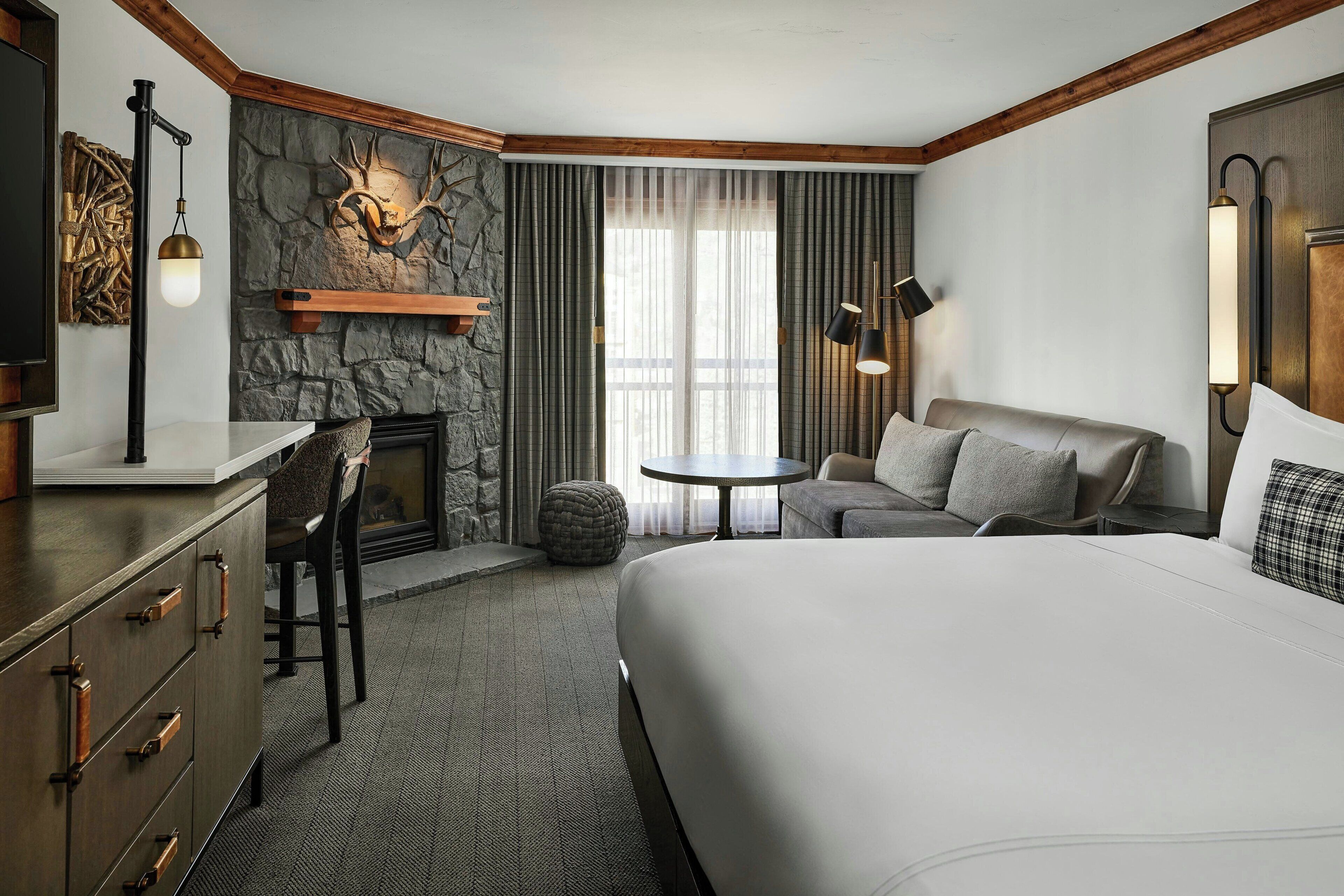 A hotel room with a large bed, a desk with a chair, a fireplace, a sofa, and a small round table.