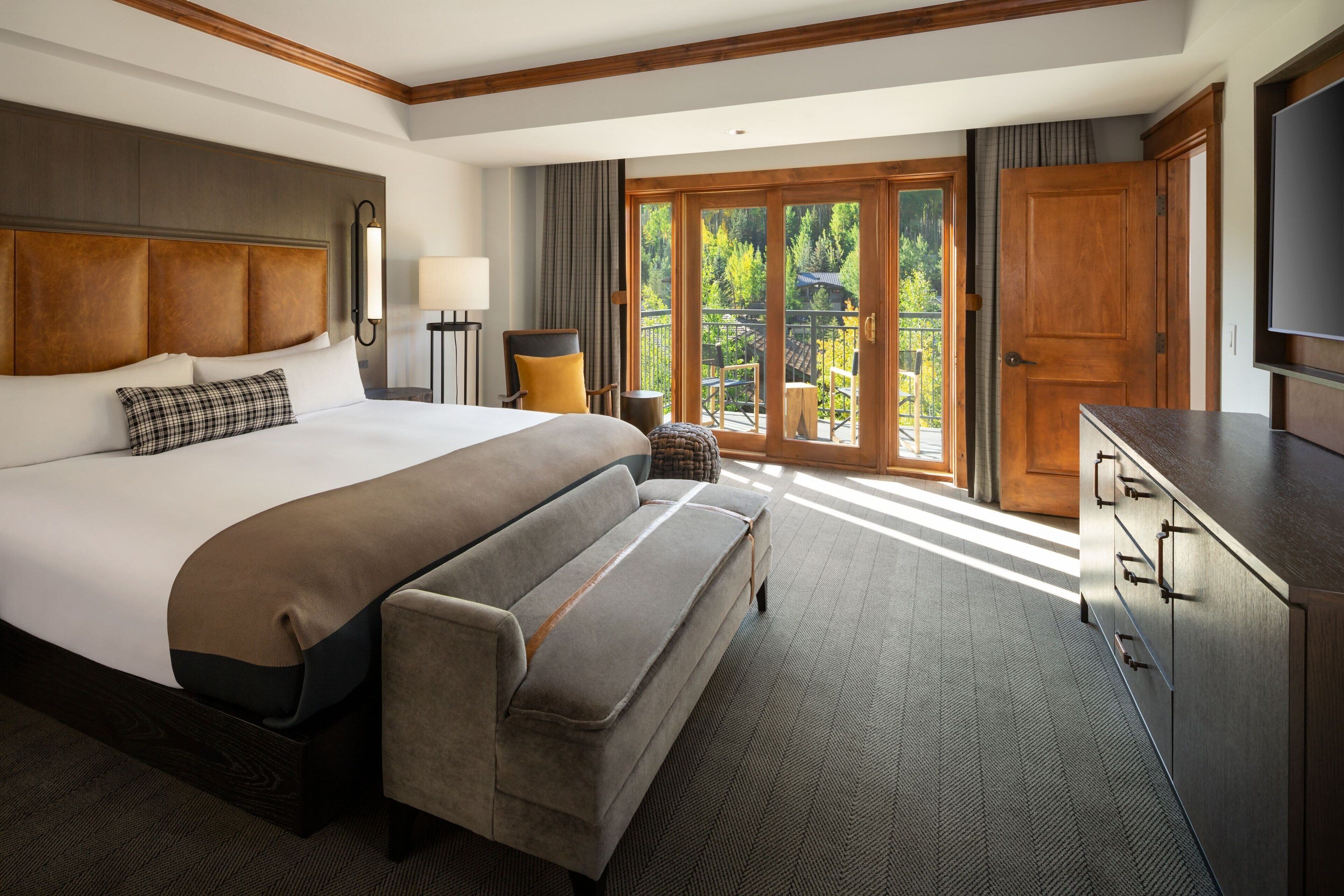 A hotel room with a large bed, a sofa, a desk, and a balcony with a view of trees.