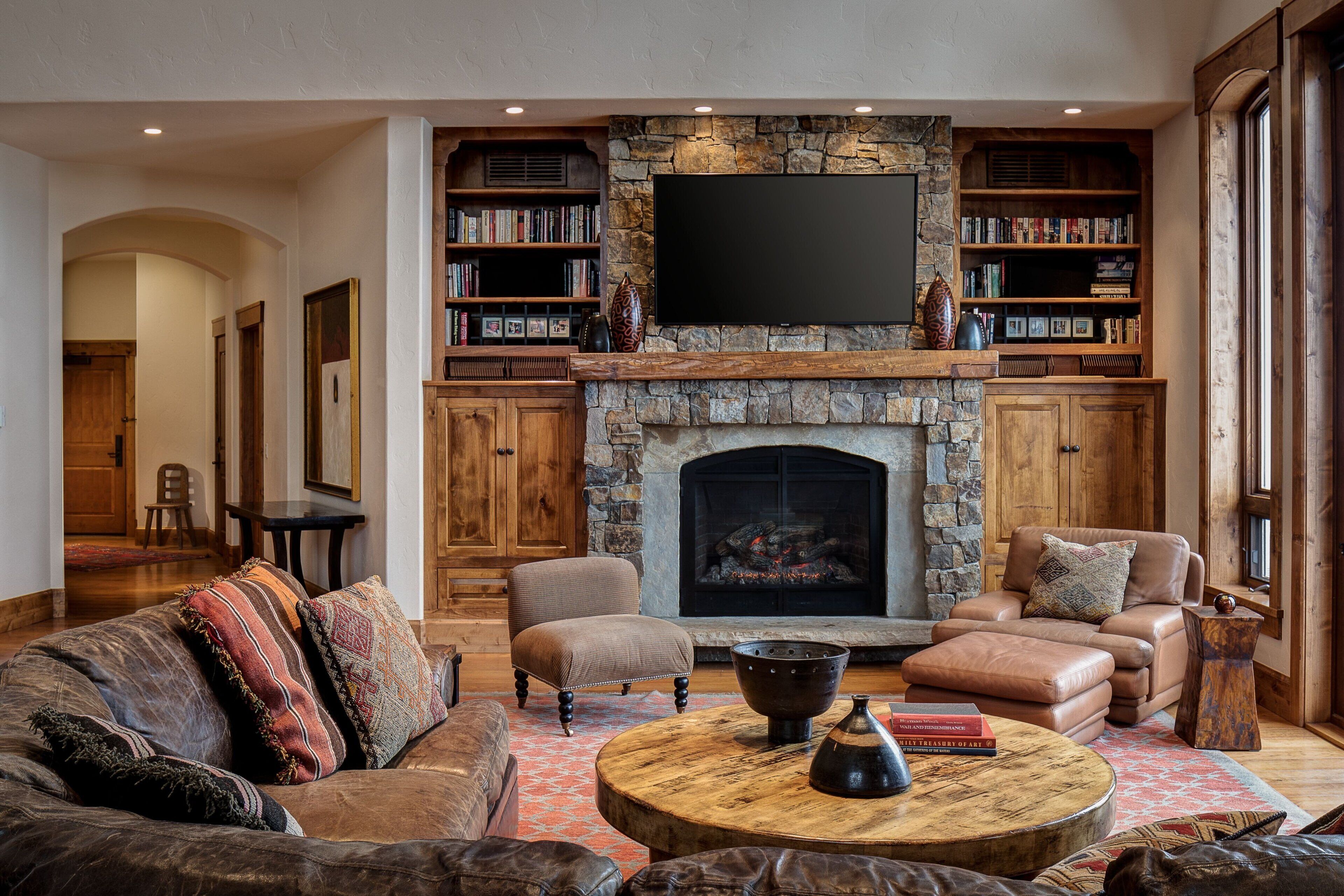 A cozy living room with a stone fireplace, leather sofas, a coffee table, and bookshelves filled with books.