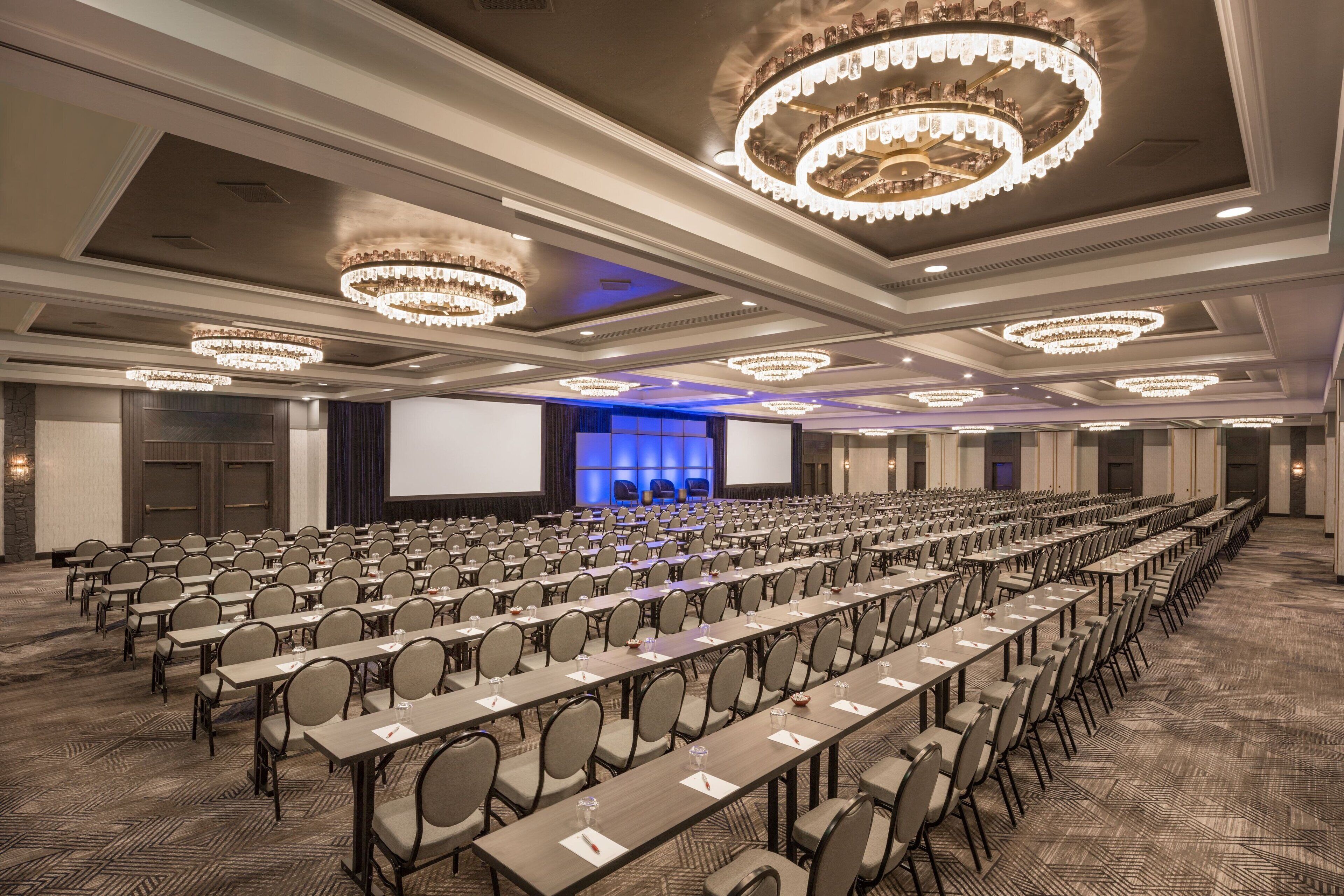 A conference room with rows of tables and chairs, large chandeliers, and a stage with a blue backdrop.