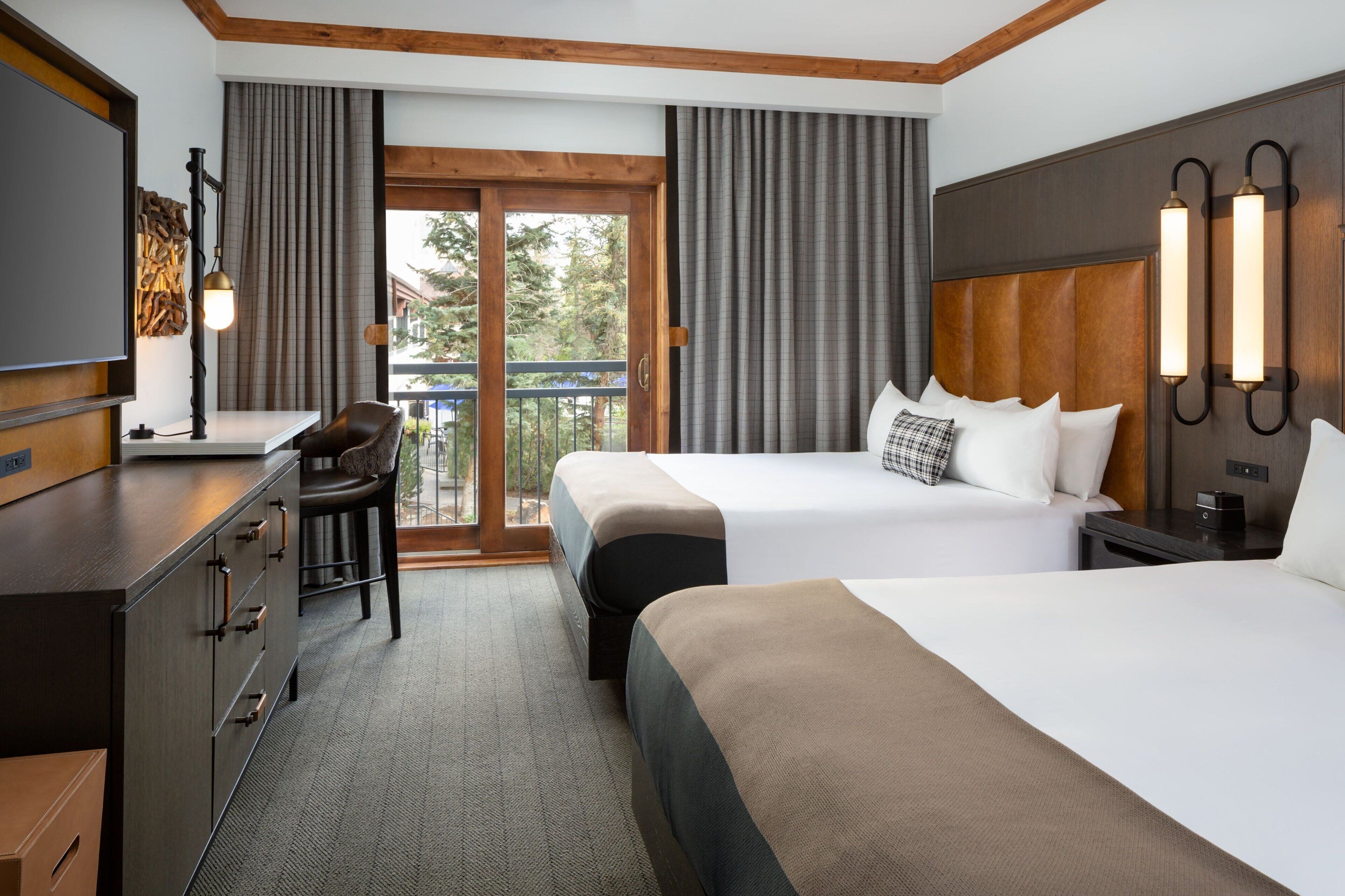 A hotel room with two beds, a desk, a chair, a TV, and a balcony with curtains.