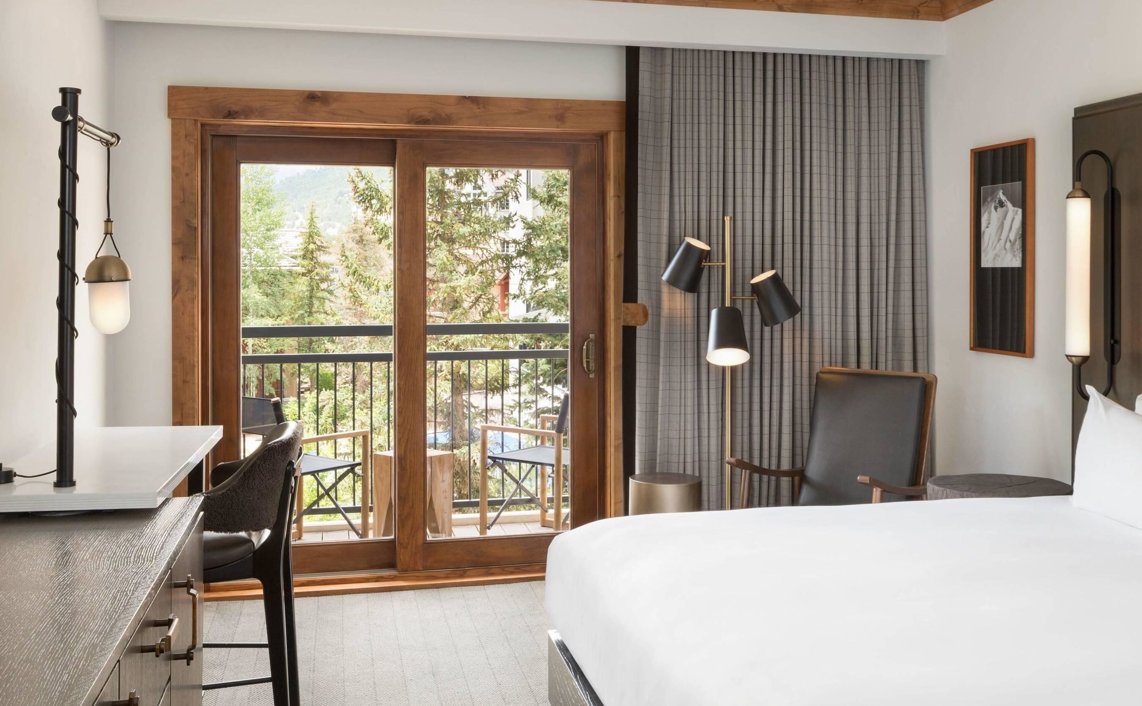 A hotel room with a bed, a desk, a chair, a balcony with a view of trees, and a large window.