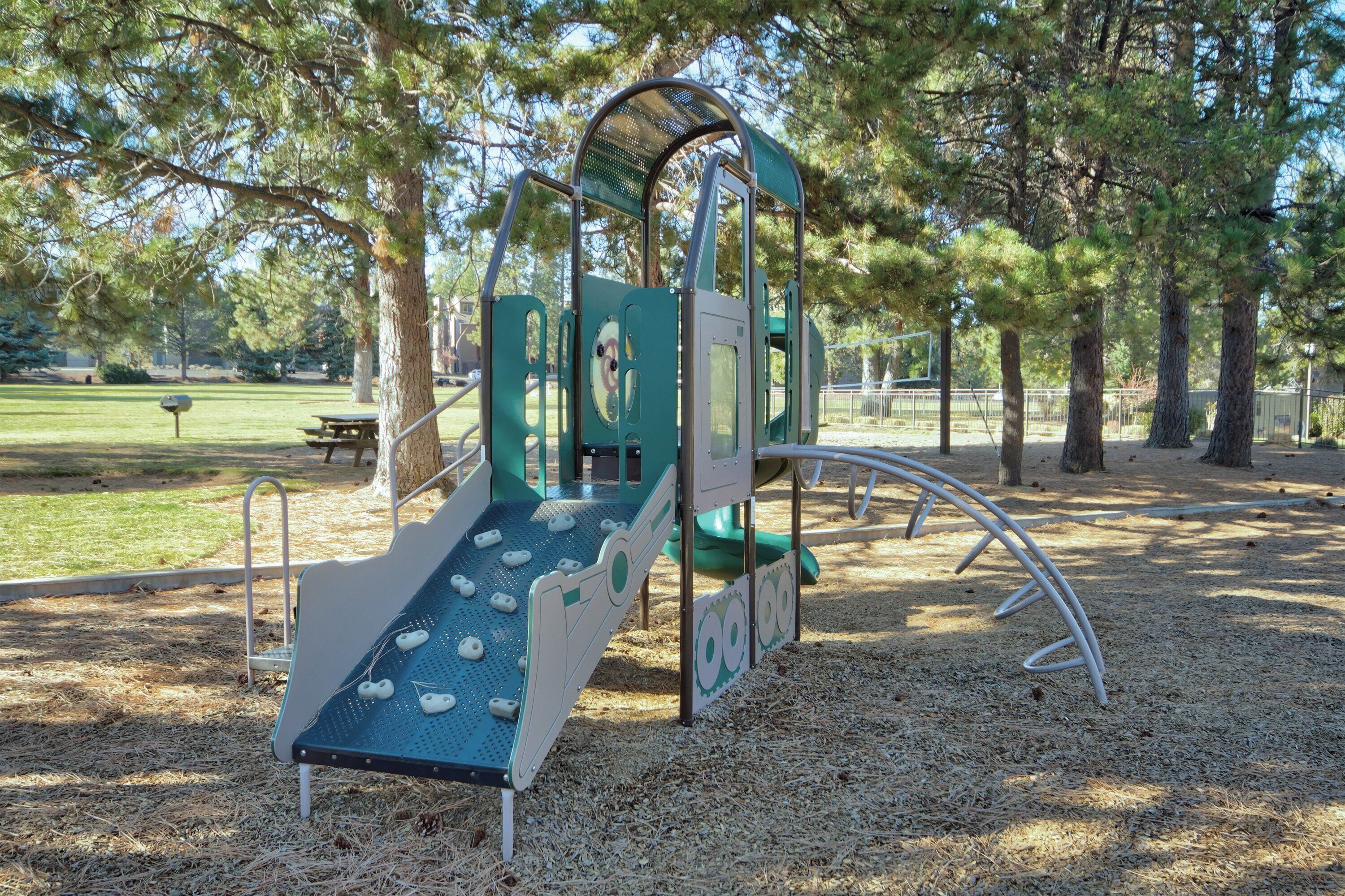 A playground with a slide and climbing structures, surrounded by trees and a picnic table in a park.