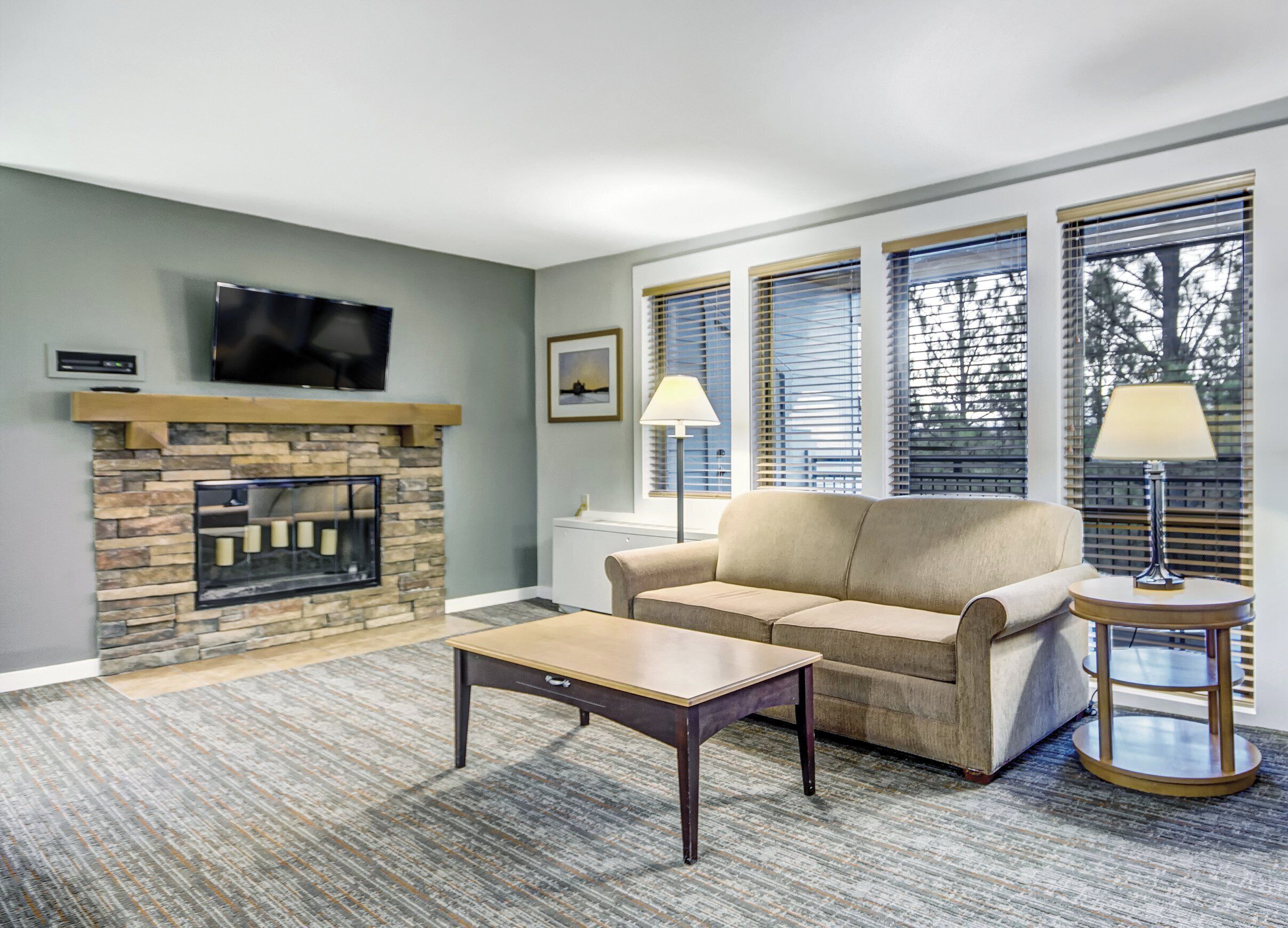 A living room with a stone fireplace, a flat-screen TV mounted above it, a beige sofa, a coffee table, and a lamp on a side table.