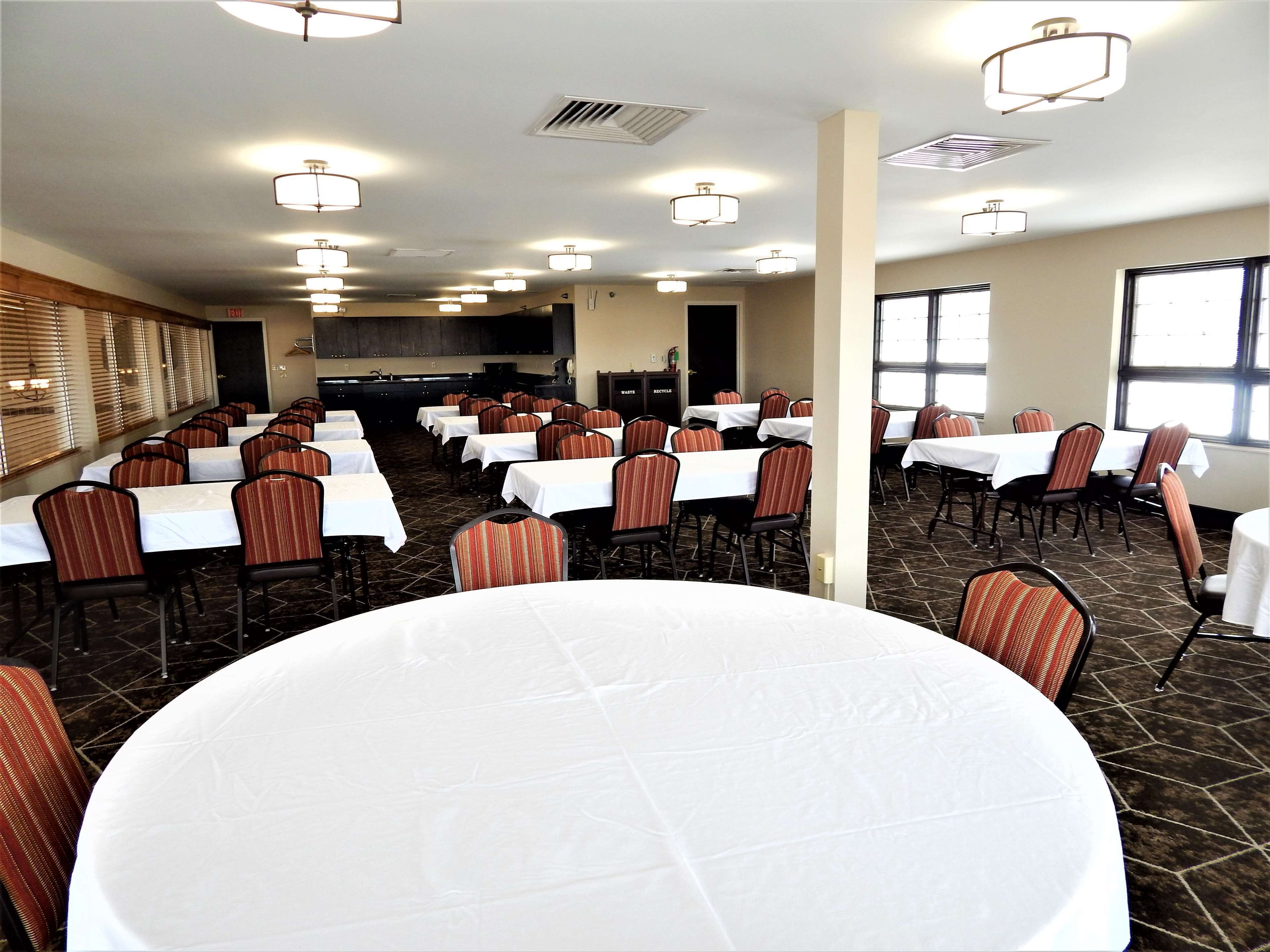 A large dining hall with multiple tables and chairs, a kitchen area in the background, and a clock on the wall.