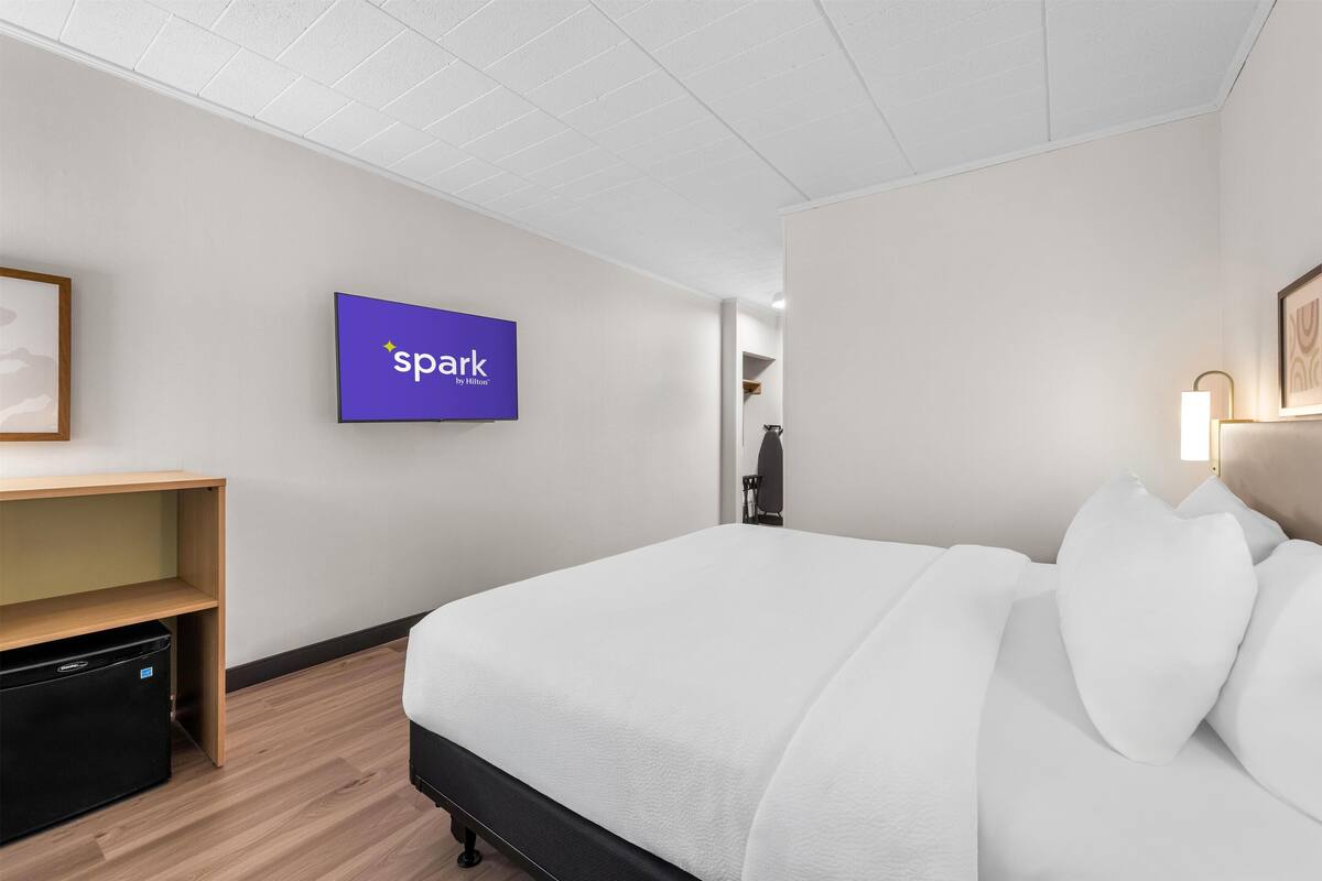 A hotel room with a bed, a TV mounted on the wall, a desk, and a small storage unit.