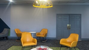 Een kamer met een moderne kroonluchter, gele stoelen, een ronde tafel met een fruitschaal, en een deur op de achtergrond.