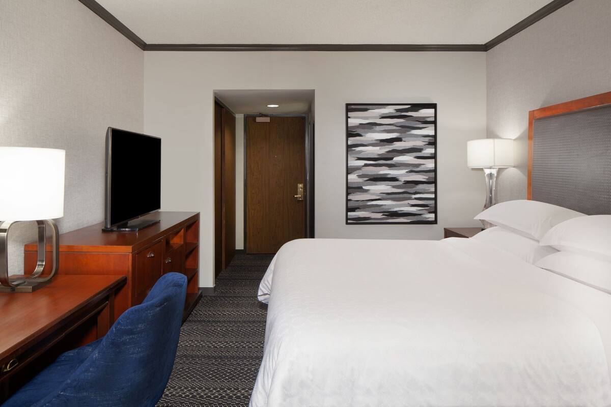 Quarto de hotel com cama, escrivaninha com televisão, cadeira, mesa de cabeceira com abajur e porta para outro aposento.