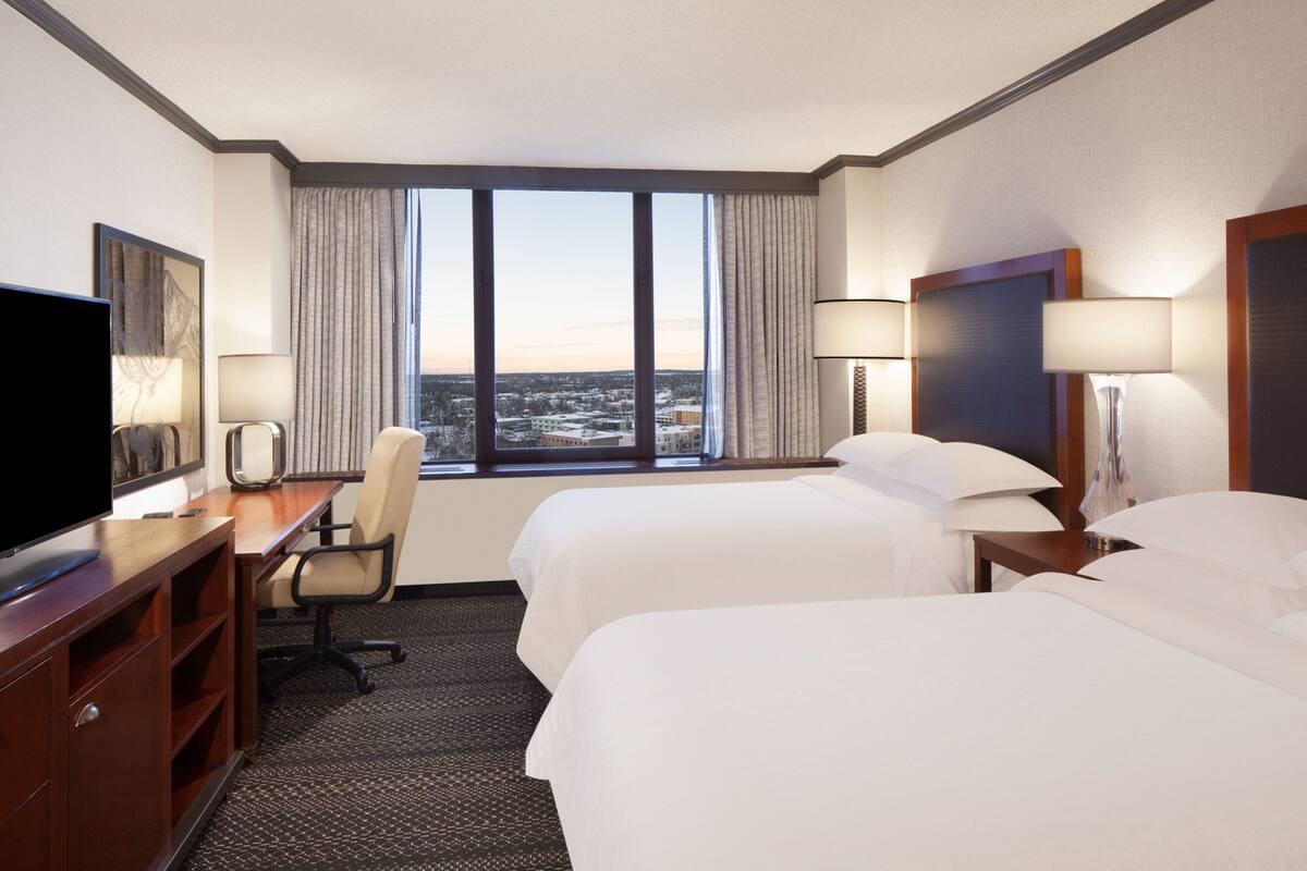 Quarto de hotel com duas camas, uma escrivaninha com televisão, uma cadeira e uma janela grande com vista para a cidade.