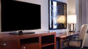 Una habitación de hotel con un escritorio de madera, un televisor, una silla, una lámpara y una obra de arte enmarcada en la pared.