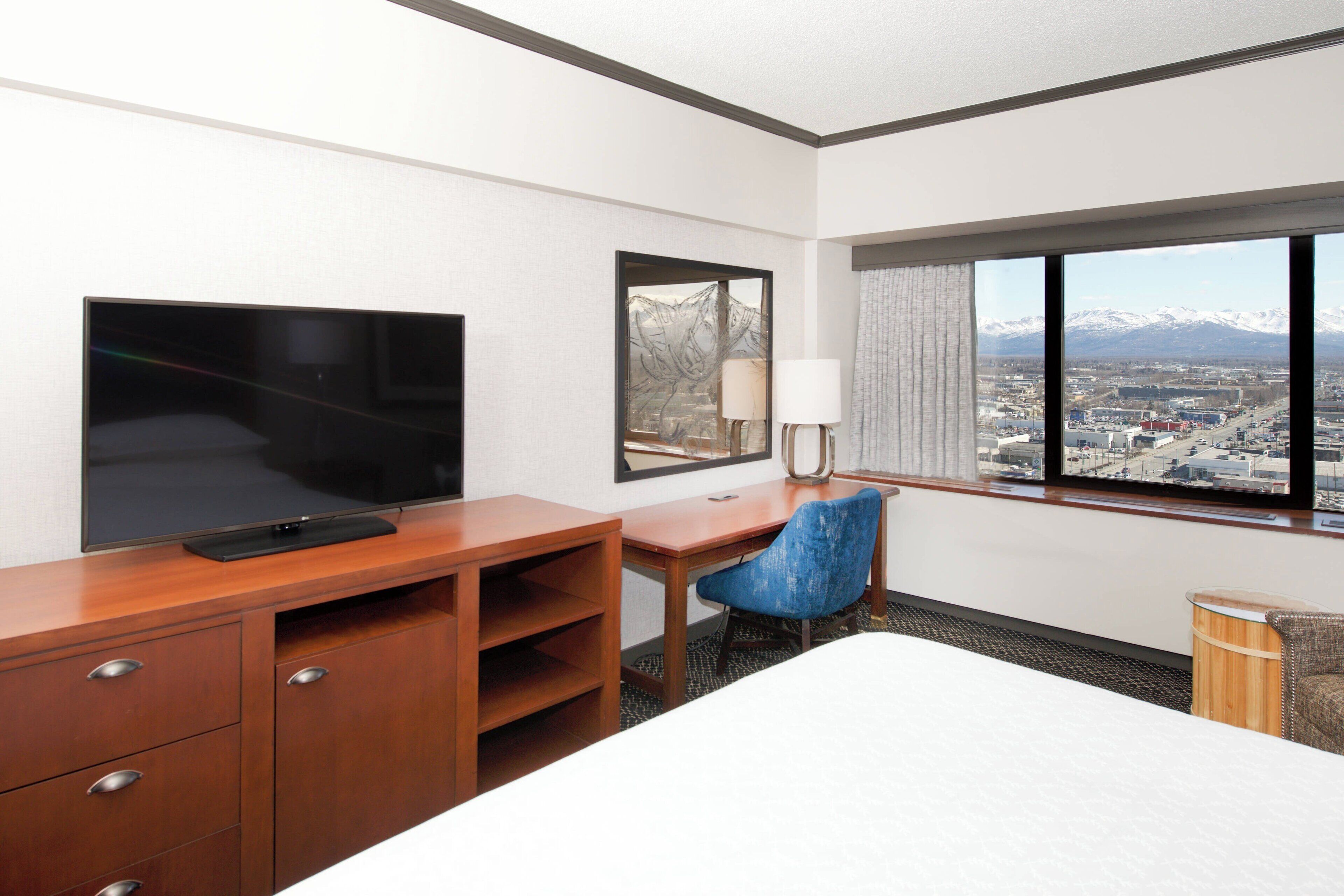 A hotel room with a bed, a desk with a chair, a TV, a window with a view of mountains, and a lamp.
