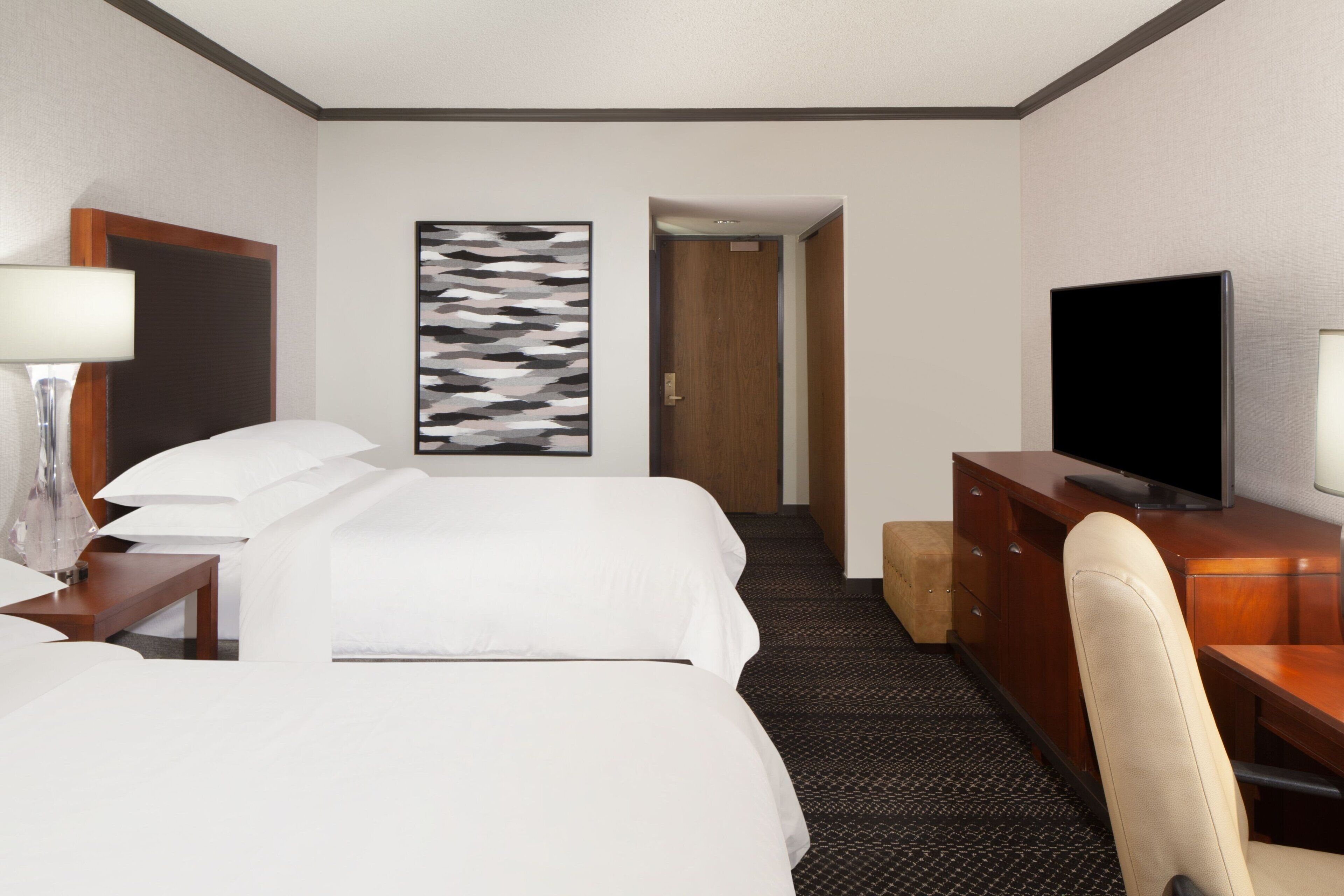 Una habitación de hotel con una cama, un escritorio con televisor, una silla, una lámpara y un cuadro en la pared.